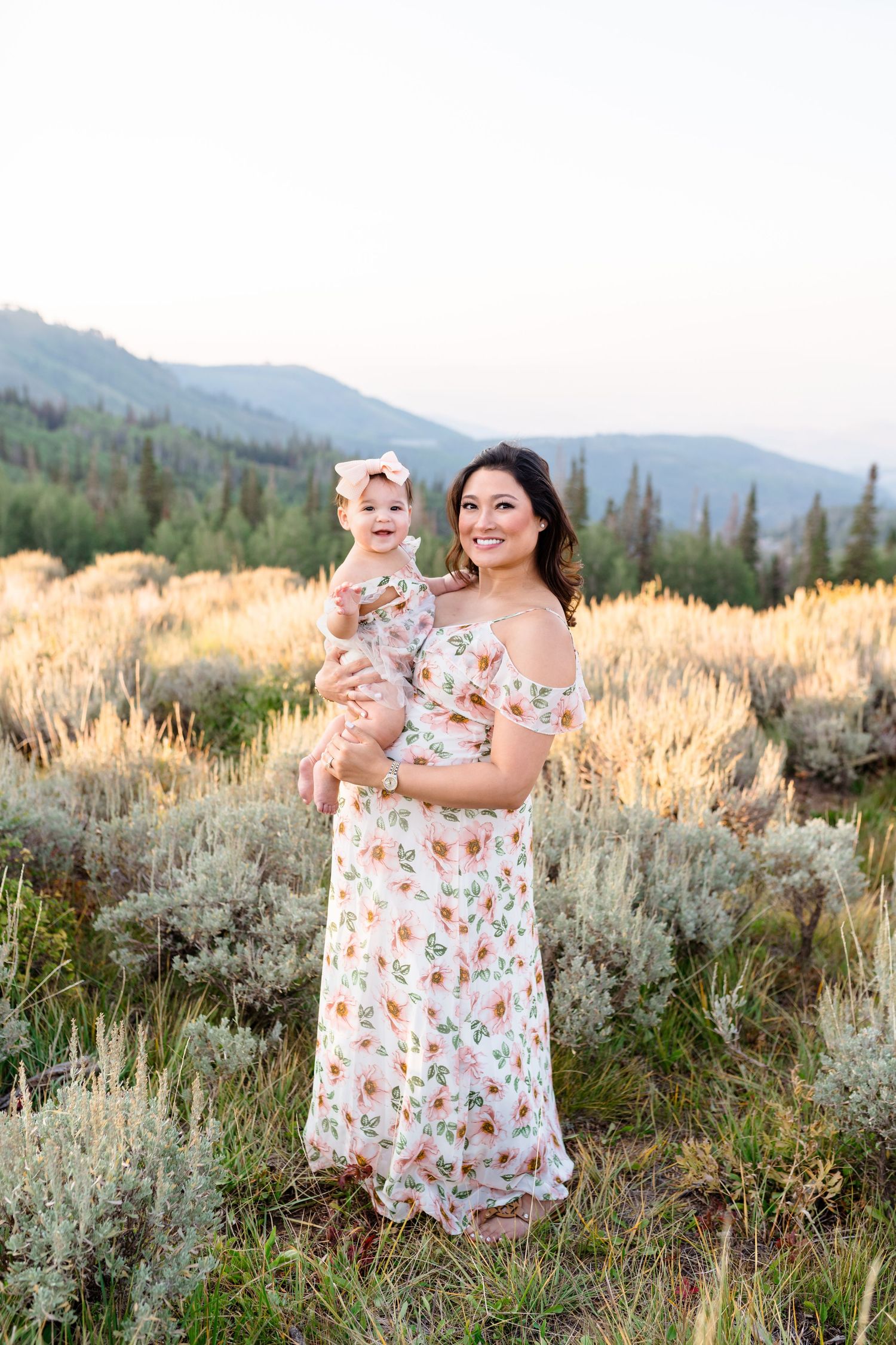 Mother and baby wearing matching floral dresses pose together in a mountain meadow at sunset.