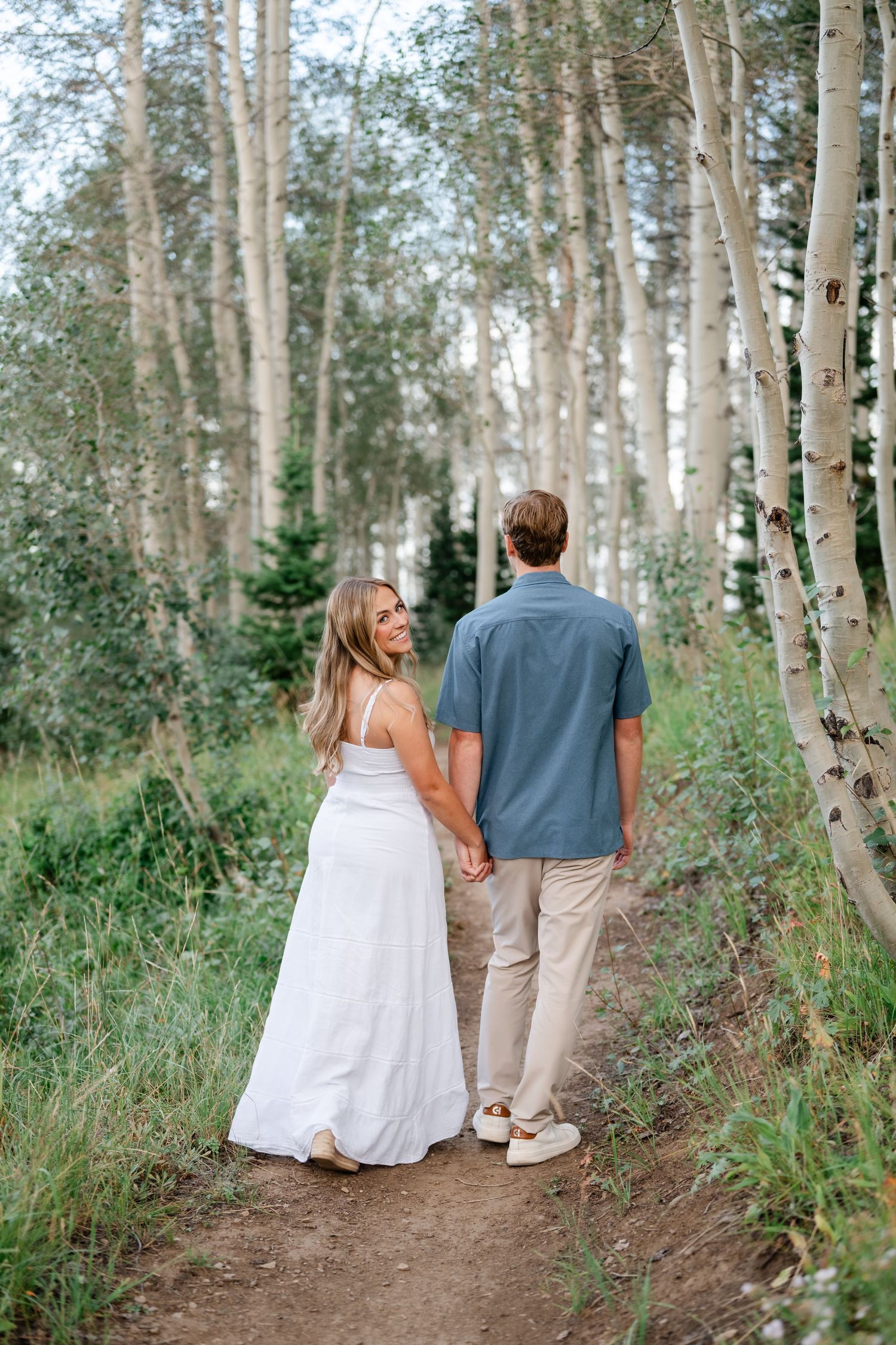 Couple holding hands walking on a forest path among aspen trees, woman in white dress looking back smiling during mountain engagements.