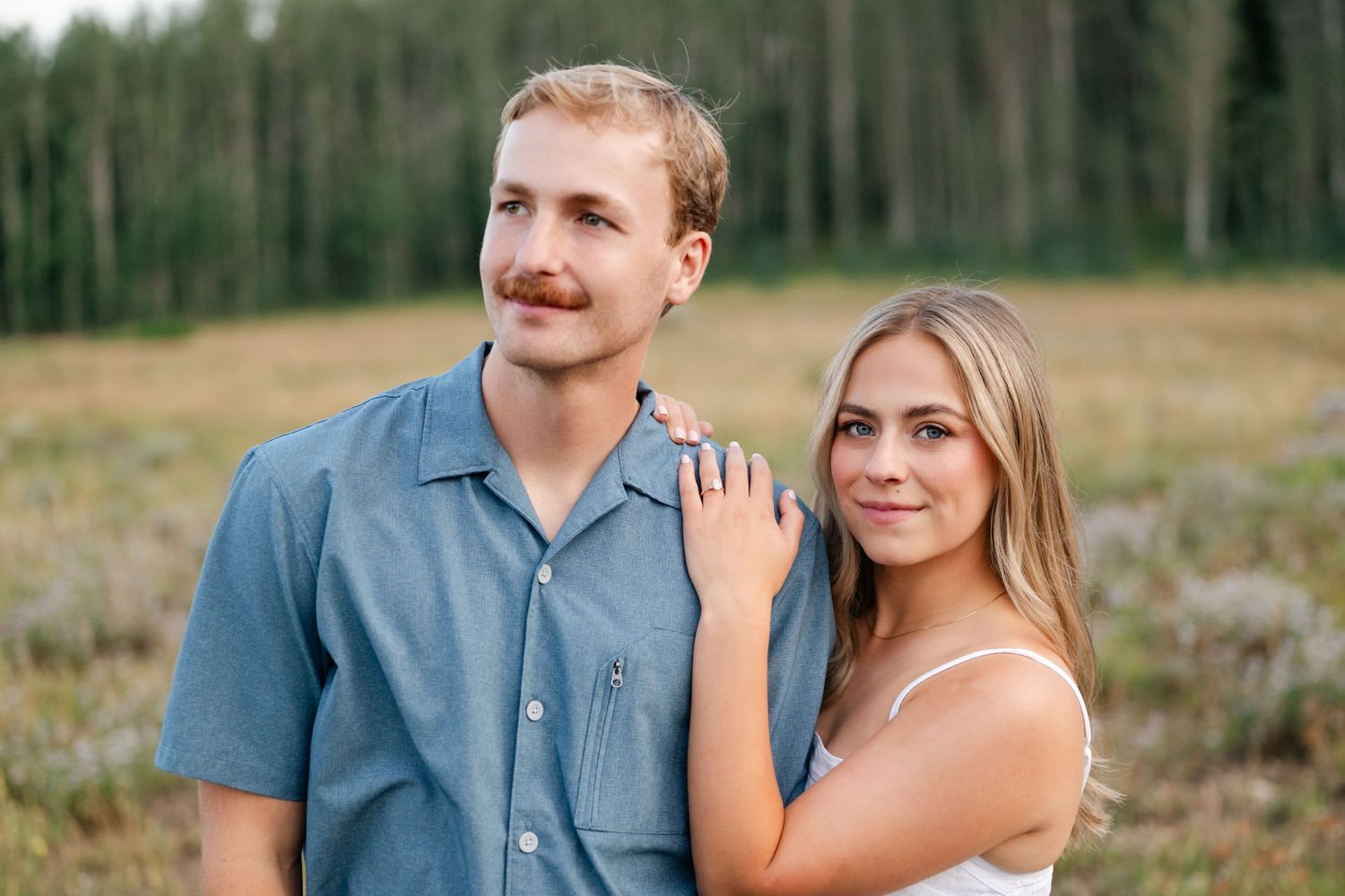 Mountain engagement photos of young couple poses outdoors in a field, man in blue shirt with mustache, woman in white top smiling warmly.