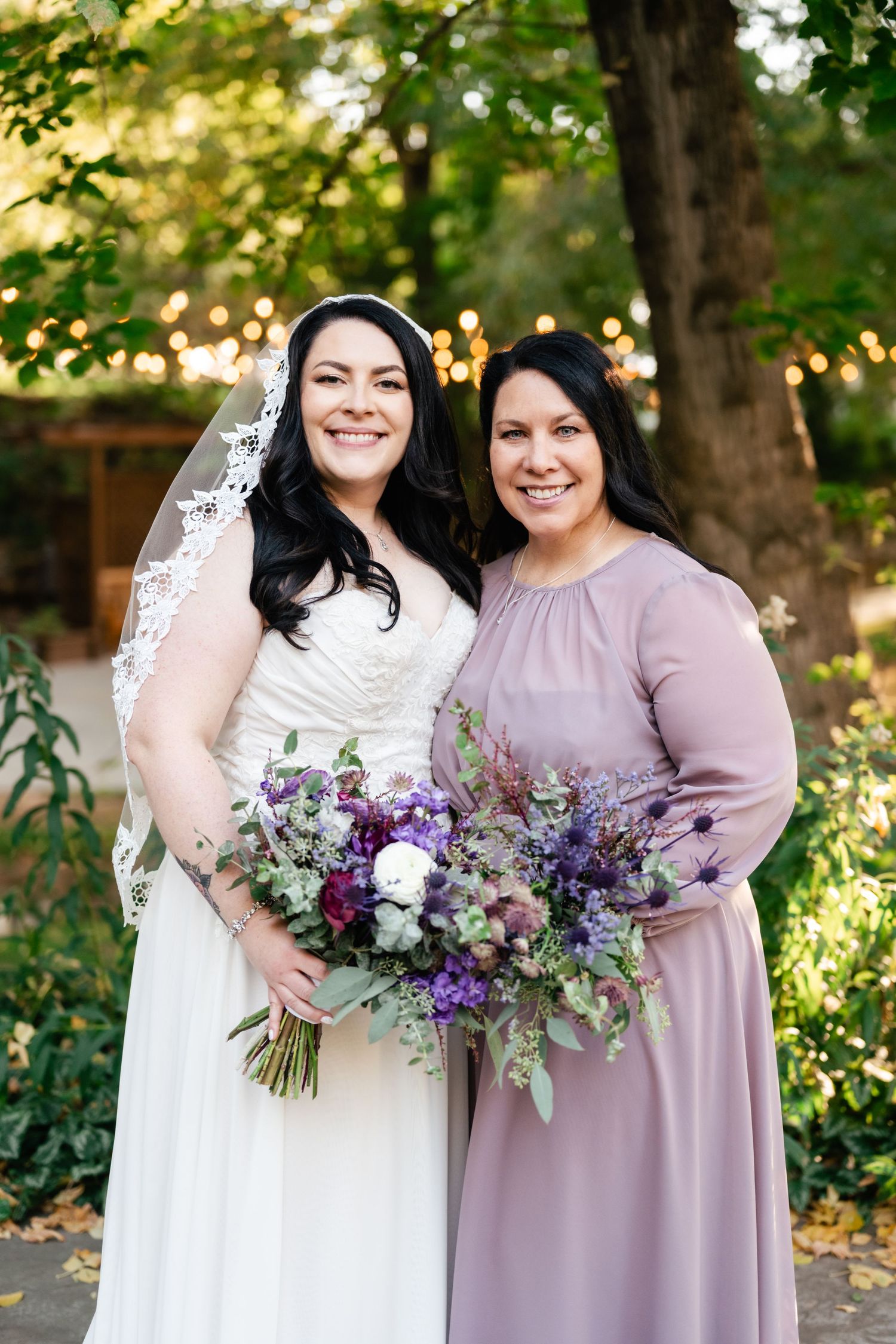 Beautiful outdoor wedding scene with bride in white lace gown and bridesmaid in mauve dress holding purple floral bouquets.