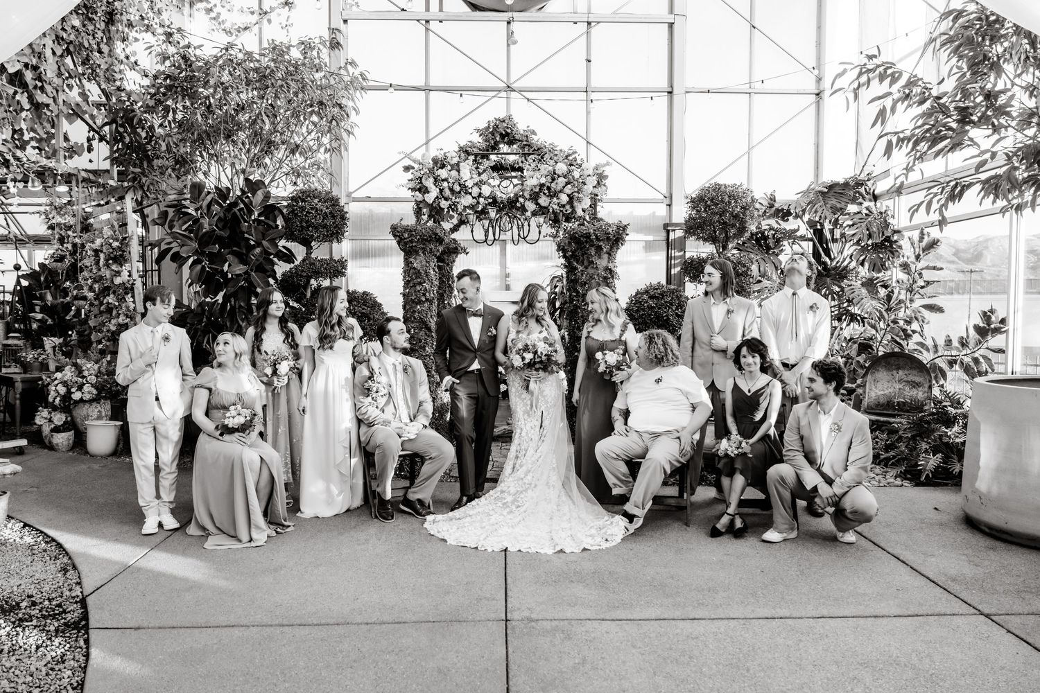 Black and white wedding party photo shows bridal group gathered under elegant greenhouse archway at indoor venue.