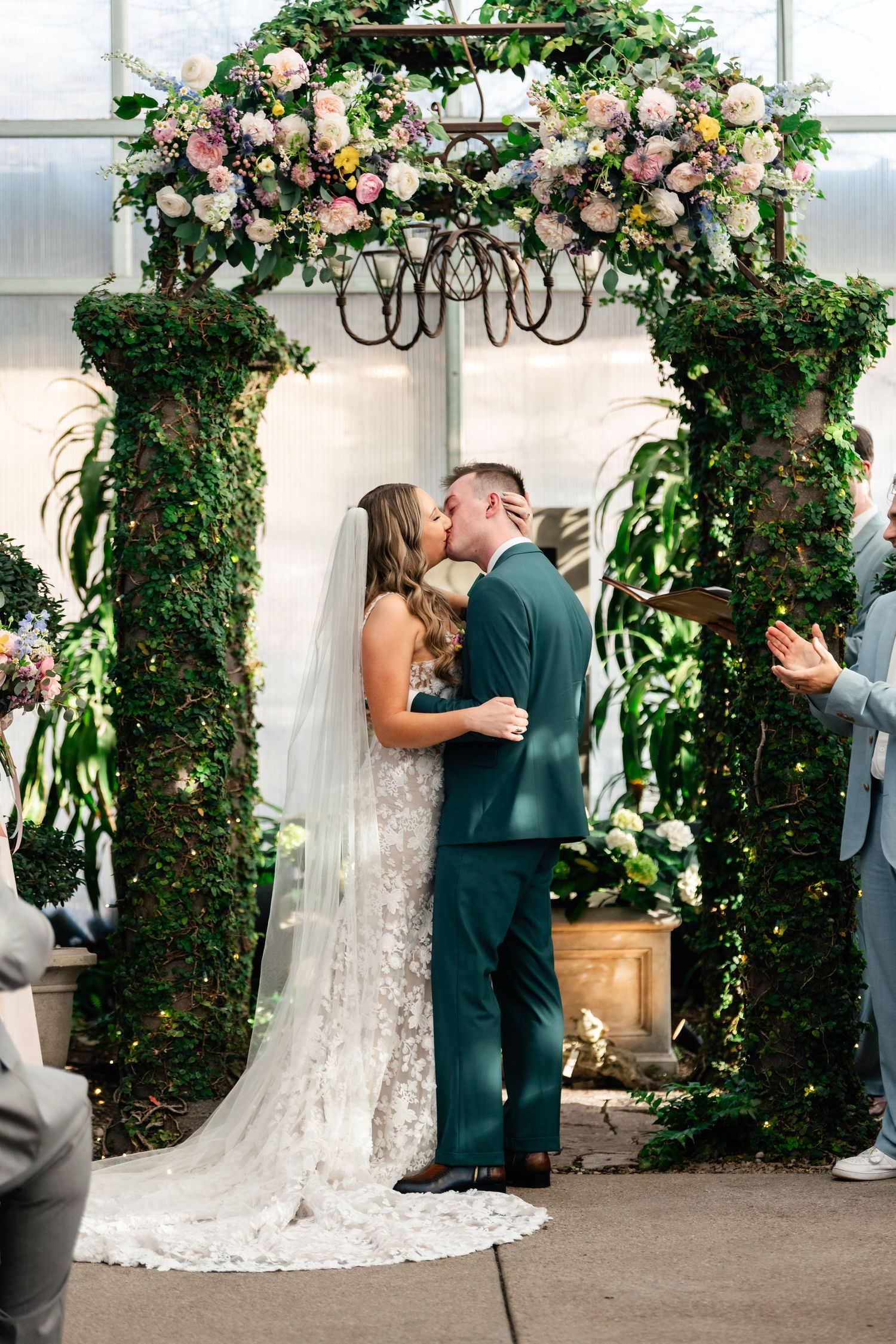 A couple shares a kiss under an elegant floral arch with pink roses and greenery at an indoor wedding ceremony.