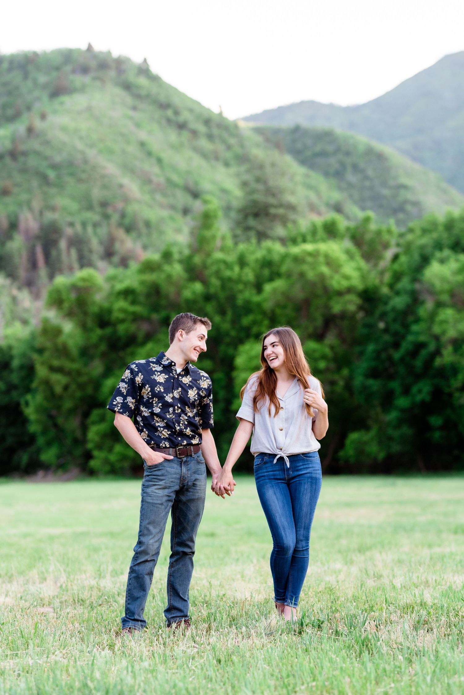 A couple holding hands while standing in a green meadow with rolling hills behind them.