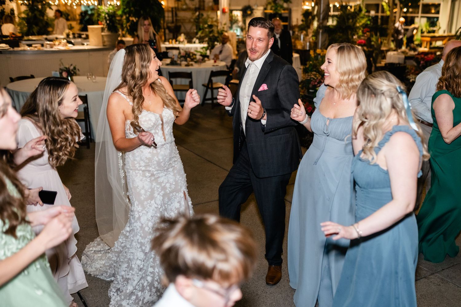Wedding guests dance and celebrate at an indoor reception venue with festive lighting.
