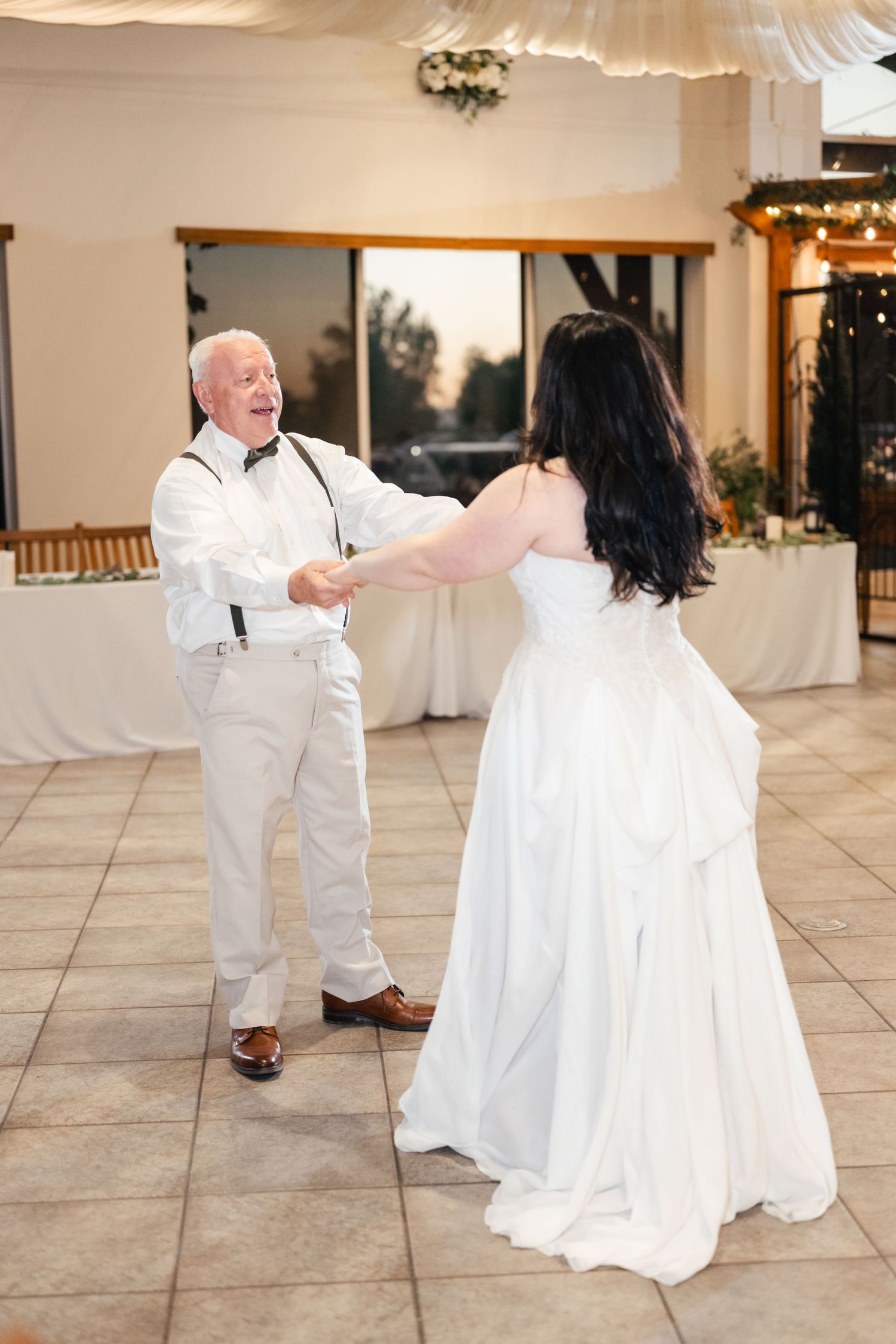 A father-daughter dance takes place in an elegant reception hall decorated with white drapes and twinkling lights.