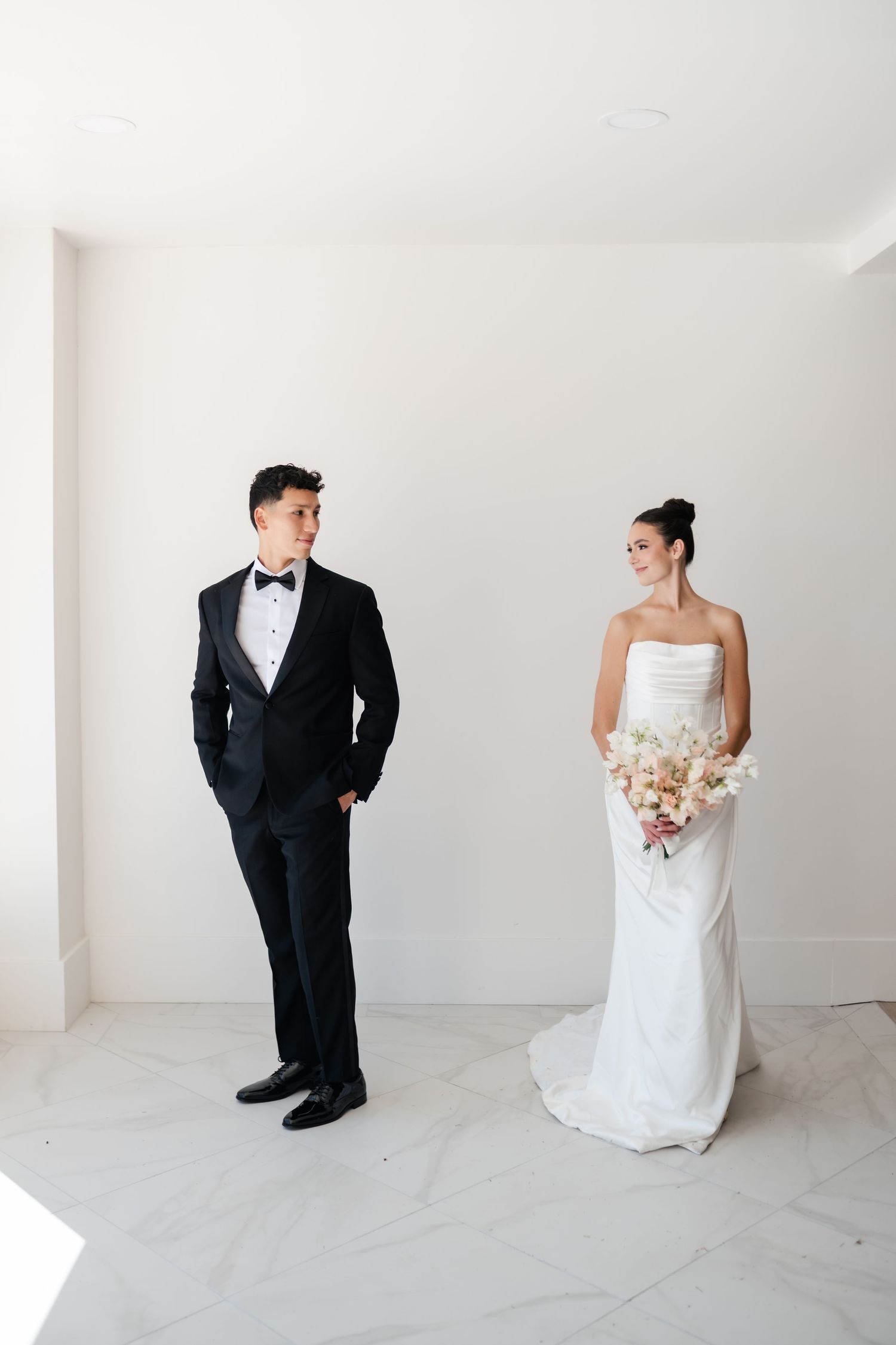A couple in formal wedding attire stand apart in a minimalist white entry of Moments on Main, bride holding a soft-colored bouquet.