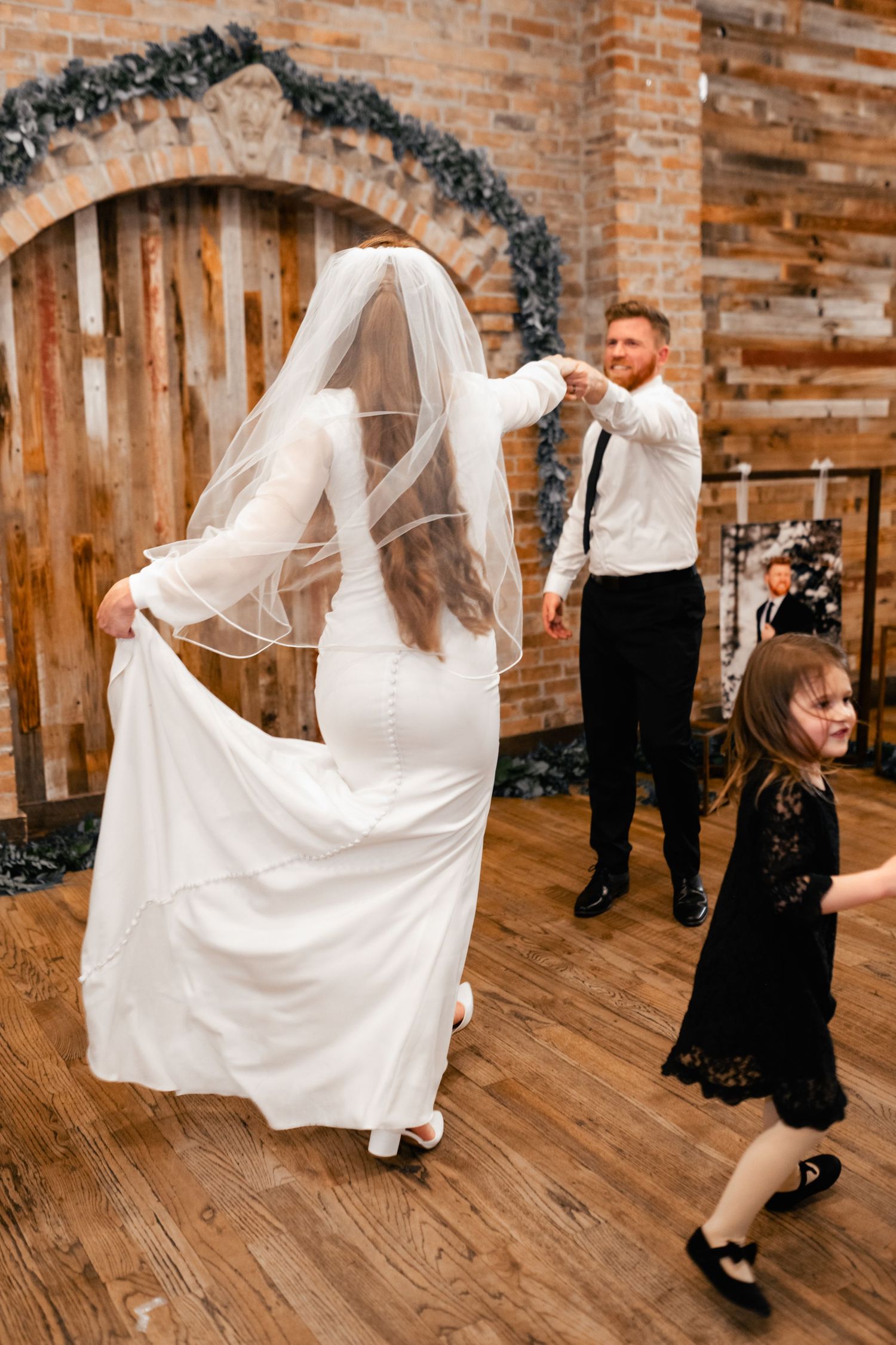 Wedding guests dance and celebrate on a wooden floor with rustic brick walls and decorative garlands in the background.
