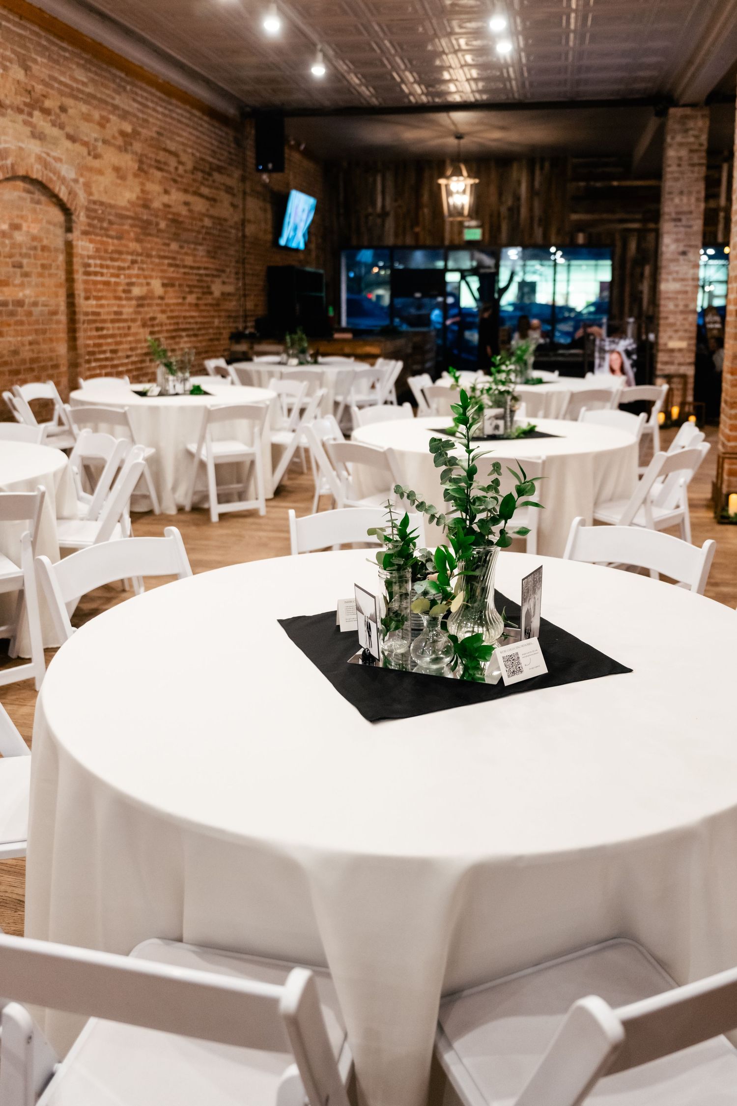 Modern event space with exposed brick walls features round tables dressed in white linens and simple greenery centerpieces.