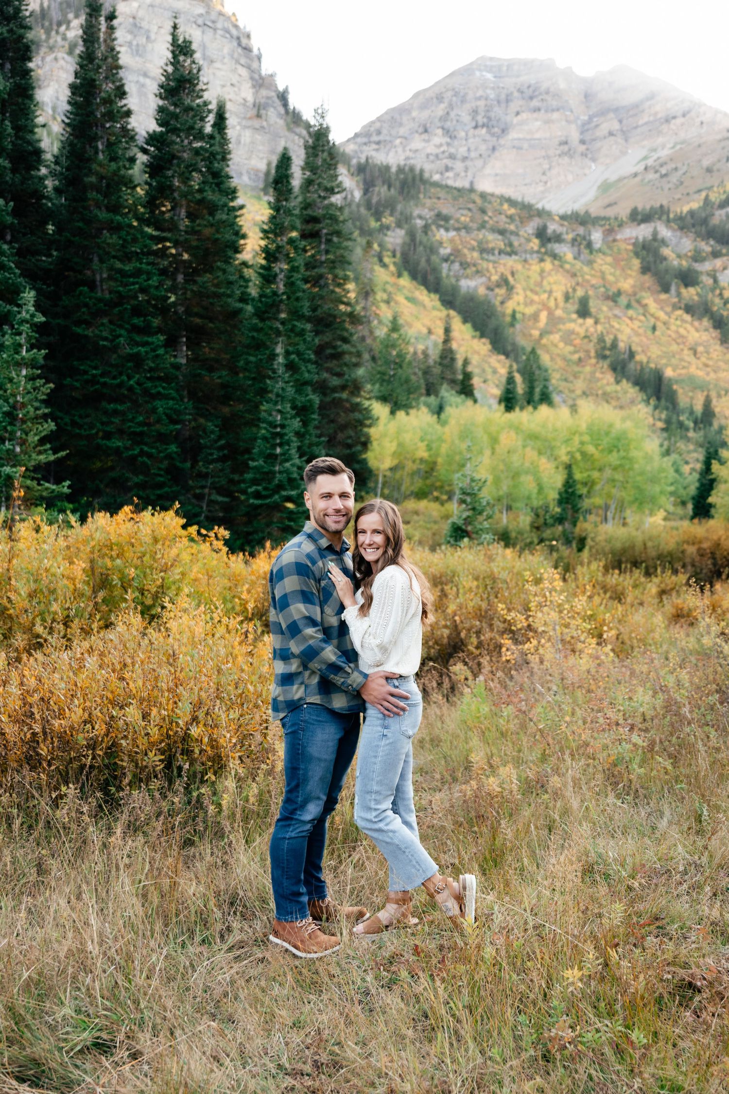 A couple poses in a scenic mountain landscape with evergreen trees and golden fall foliage in the background.