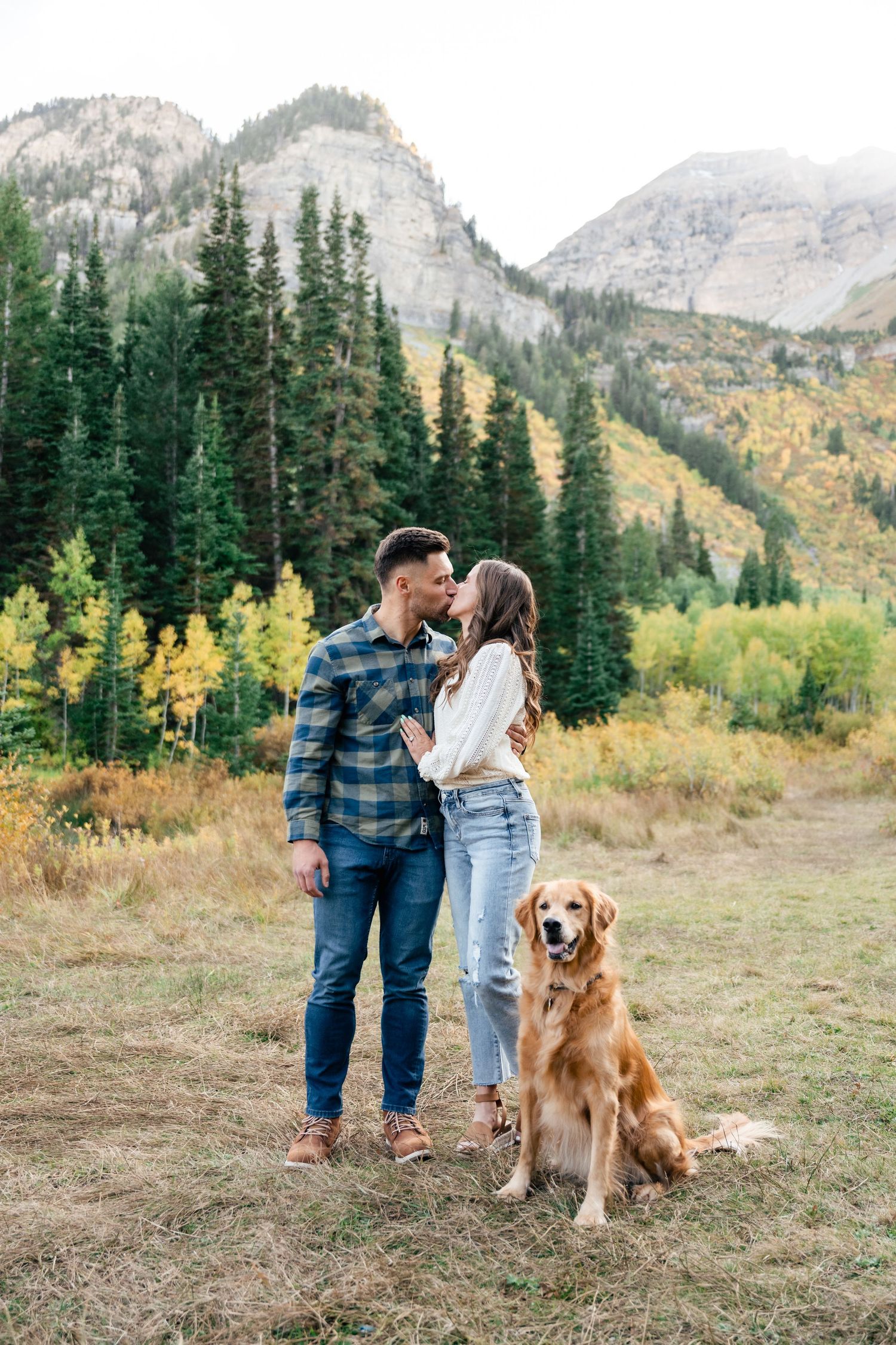 A couple and their golden retriever pose together in front of a scenic mountain landscape with fall foliage.