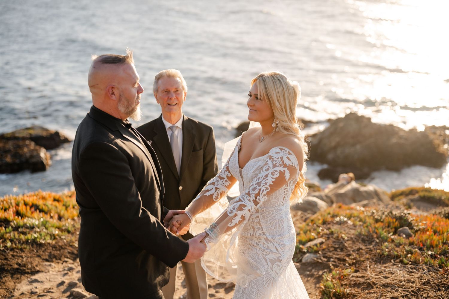A wedding ceremony takes place on a rocky coastal cliff during golden hour as waves crash against the shore below.