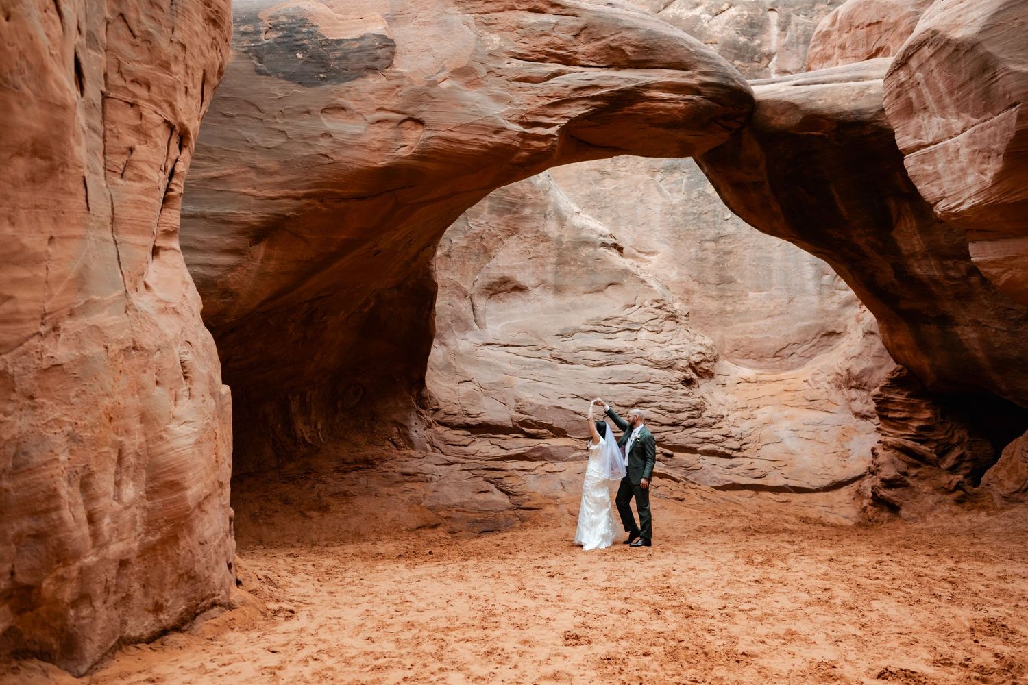A couple in wedding attire stands beneath a majestic natural stone arch in a red rock desert canyon.