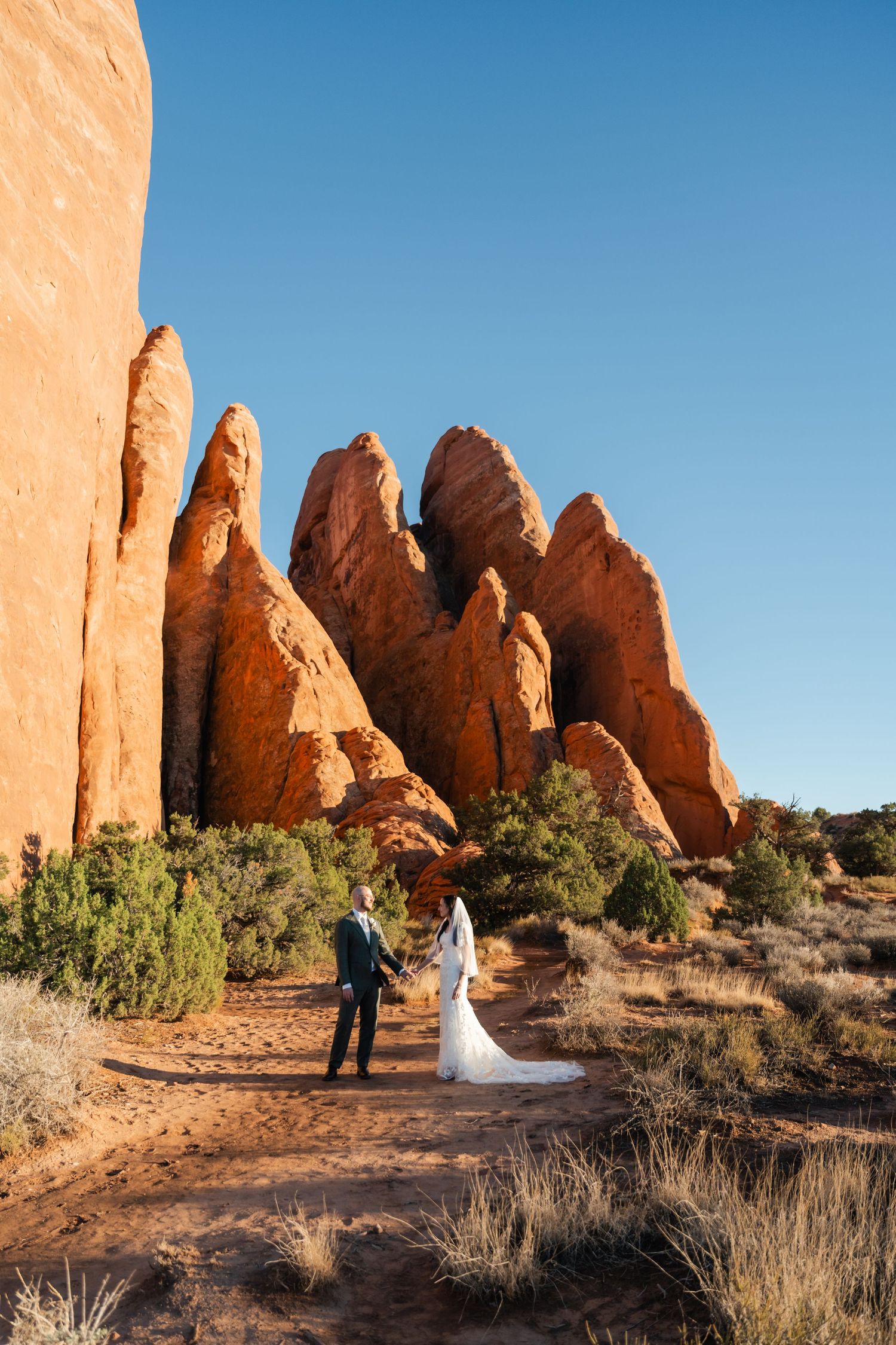 A couple in wedding attire stands against majestic red rock formations in the desert landscape.