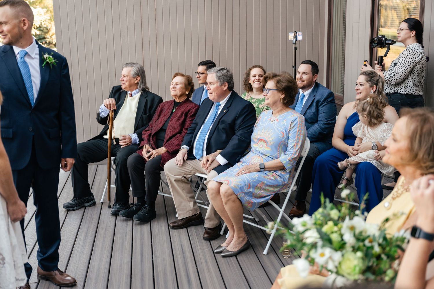 Wedding guests seated in folding chairs on an outdoor deck during a ceremony.