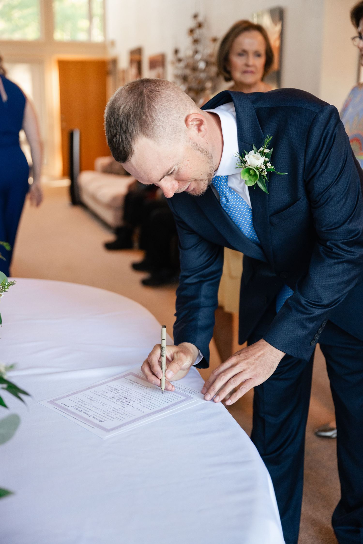 People signing a document at a formal event with guests seated in the background.