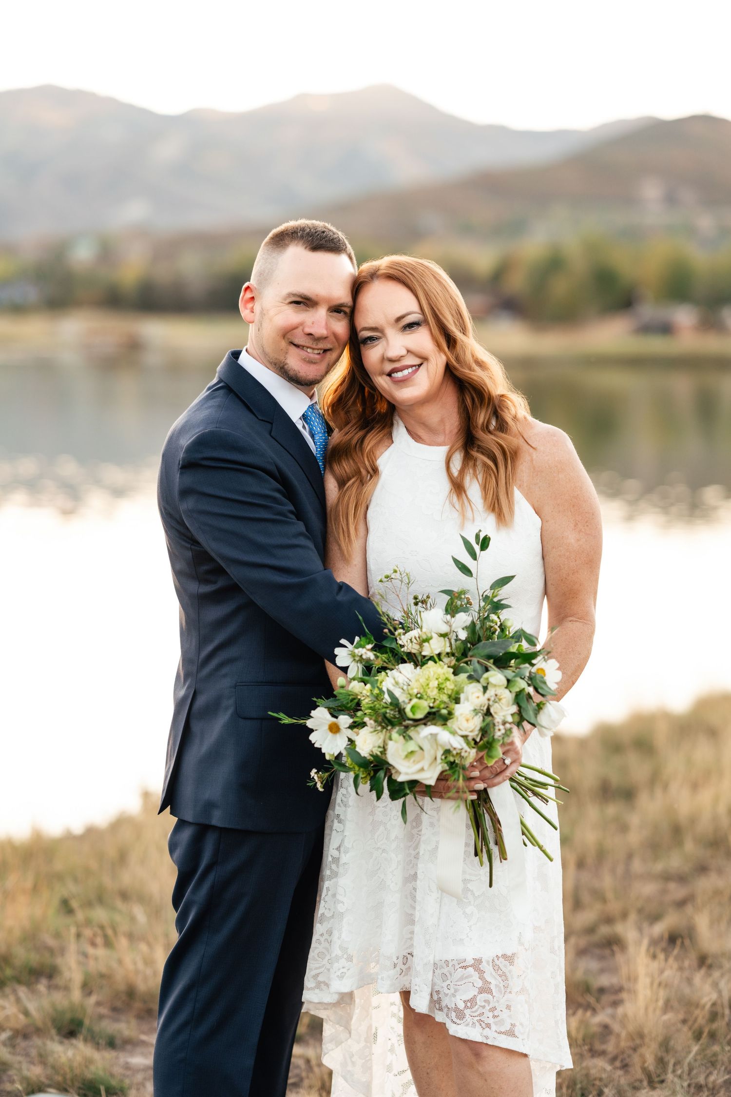 A couple embraces while holding a white and green floral bouquet near a lake with mountains in the background.