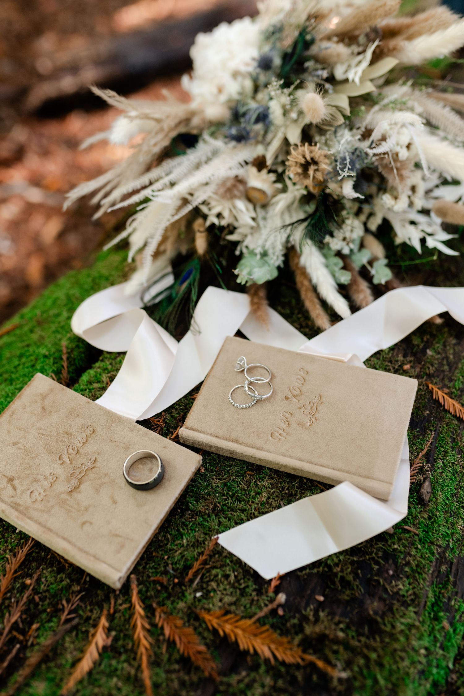 Wedding rings and vow books displayed on moss with white ribbon and dried floral bouquet.