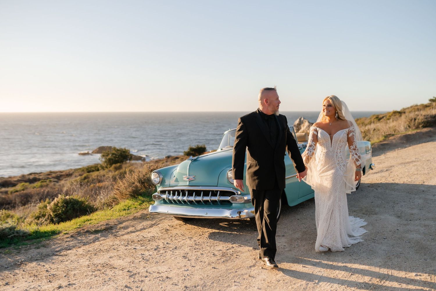 A couple poses by a classic turquoise car along a scenic coastal road.