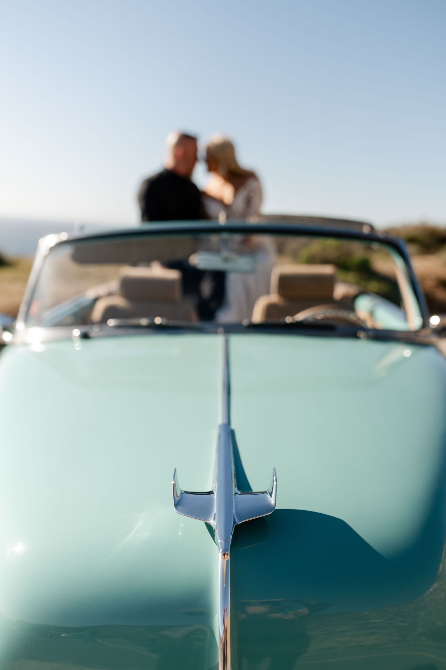 Mint green vintage convertible with hood ornament and blurred figures in background.