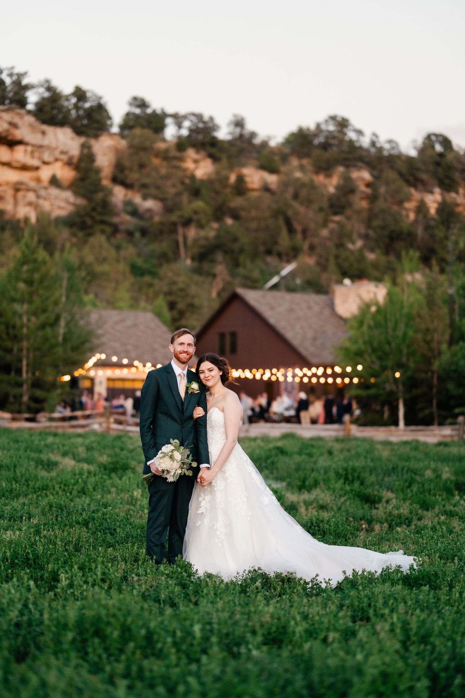 A couple in formal wedding attire stands together in green meadow with rustic buildings and string lights at dusk. Ranch house wedding.