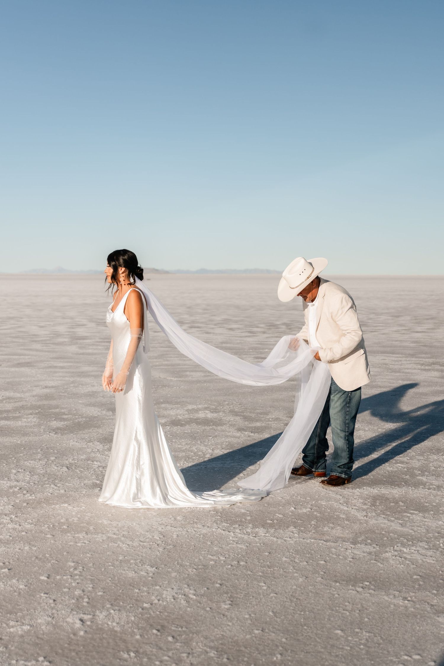 Wedding couple poses playfully with flowing white veil on salt flats during sunset photoshoot.