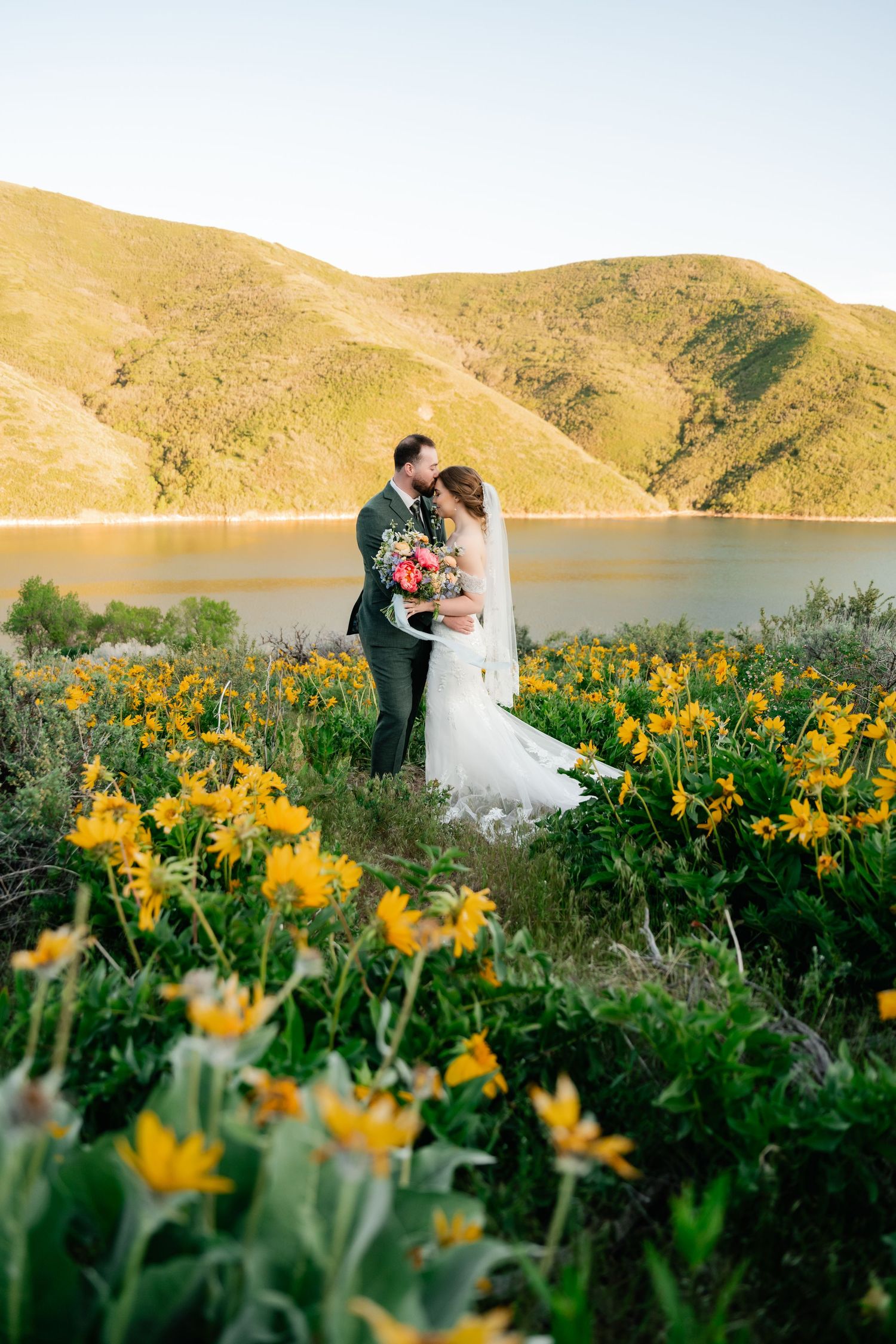 Newlyweds embrace in field of yellow wildflowers with mountain lake backdrop.