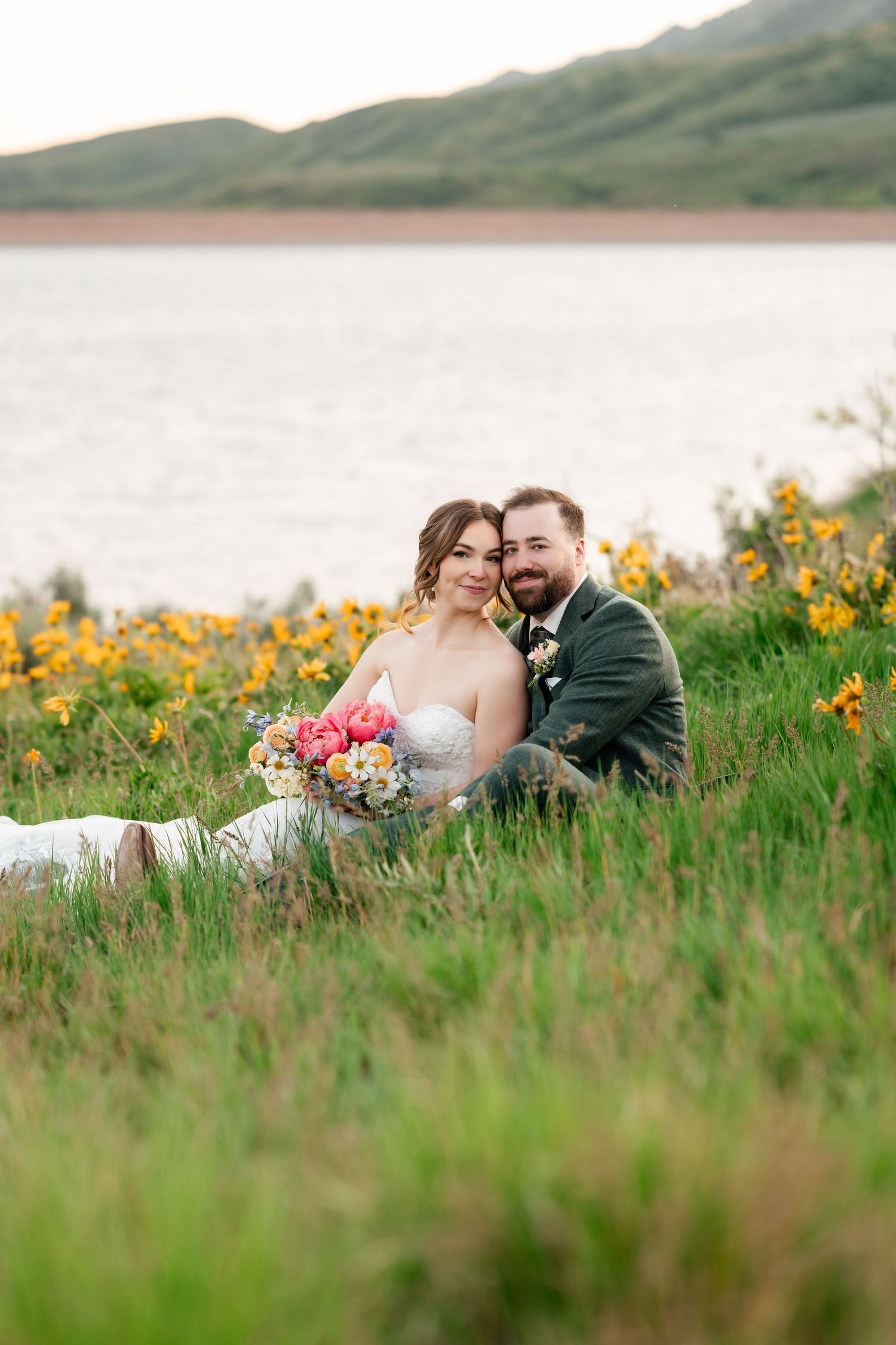 Wedding couple rests in mountain wildflower meadow near lake during sunset.