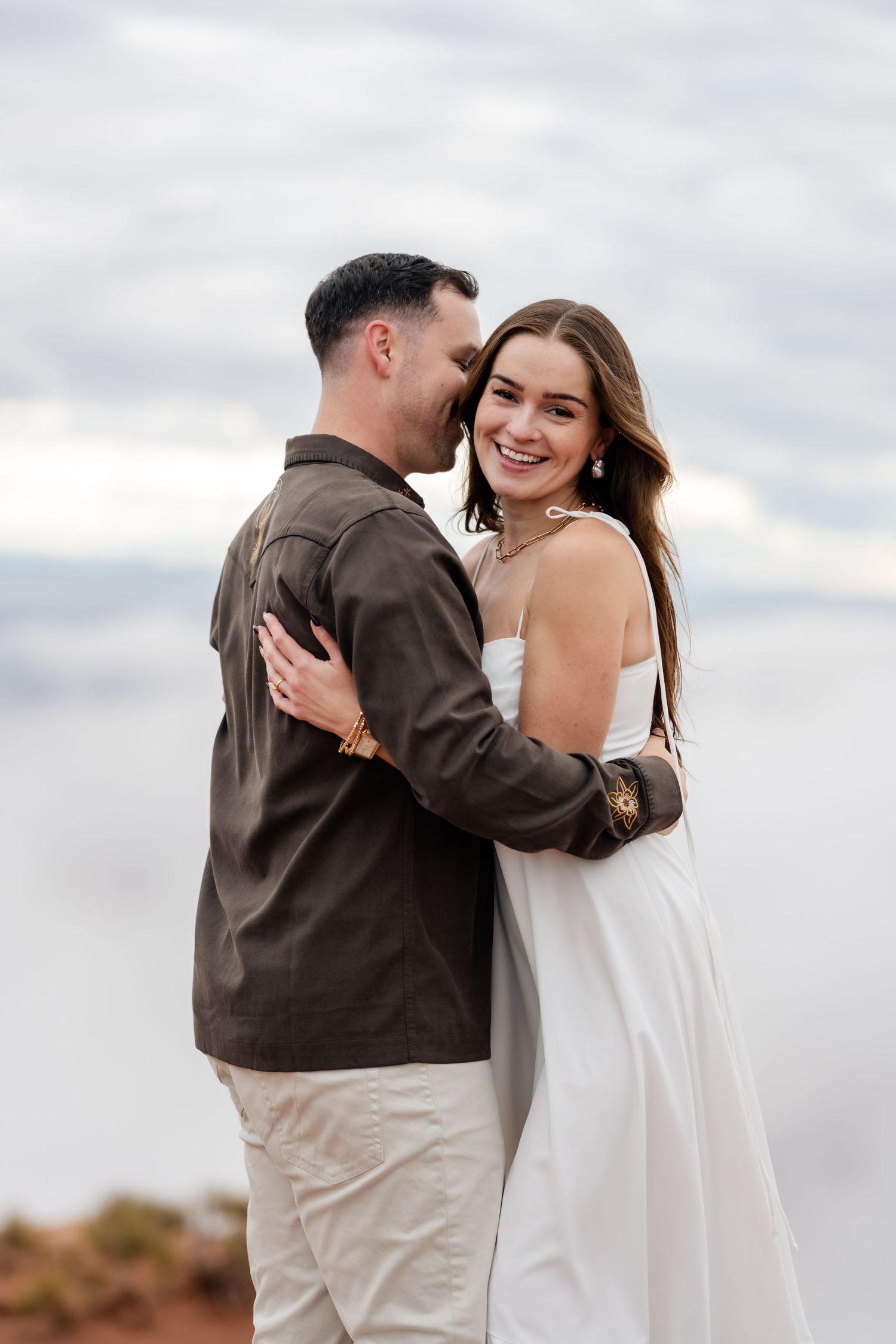 A romantic couple embraces on a cloudy day, with the person in brown jacket and khaki pants holding their partner in white.