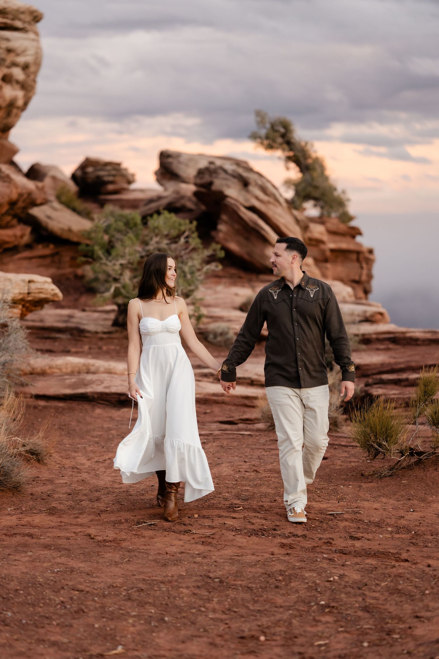 Couple walking hand in hand along red rock formations under cloudy desert skies.