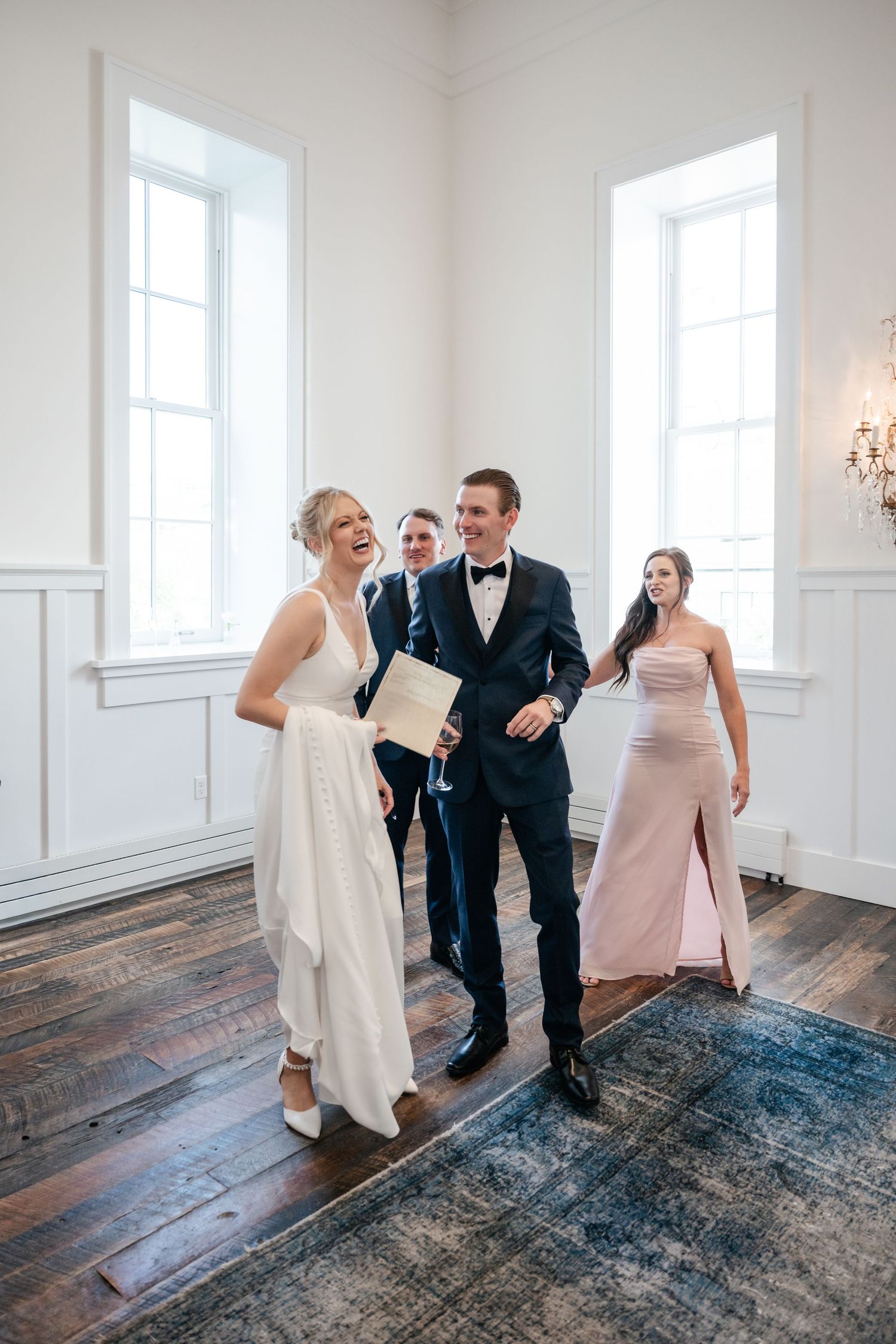 Wedding party laughs together in bright white room with hardwood floors and tall windows.
