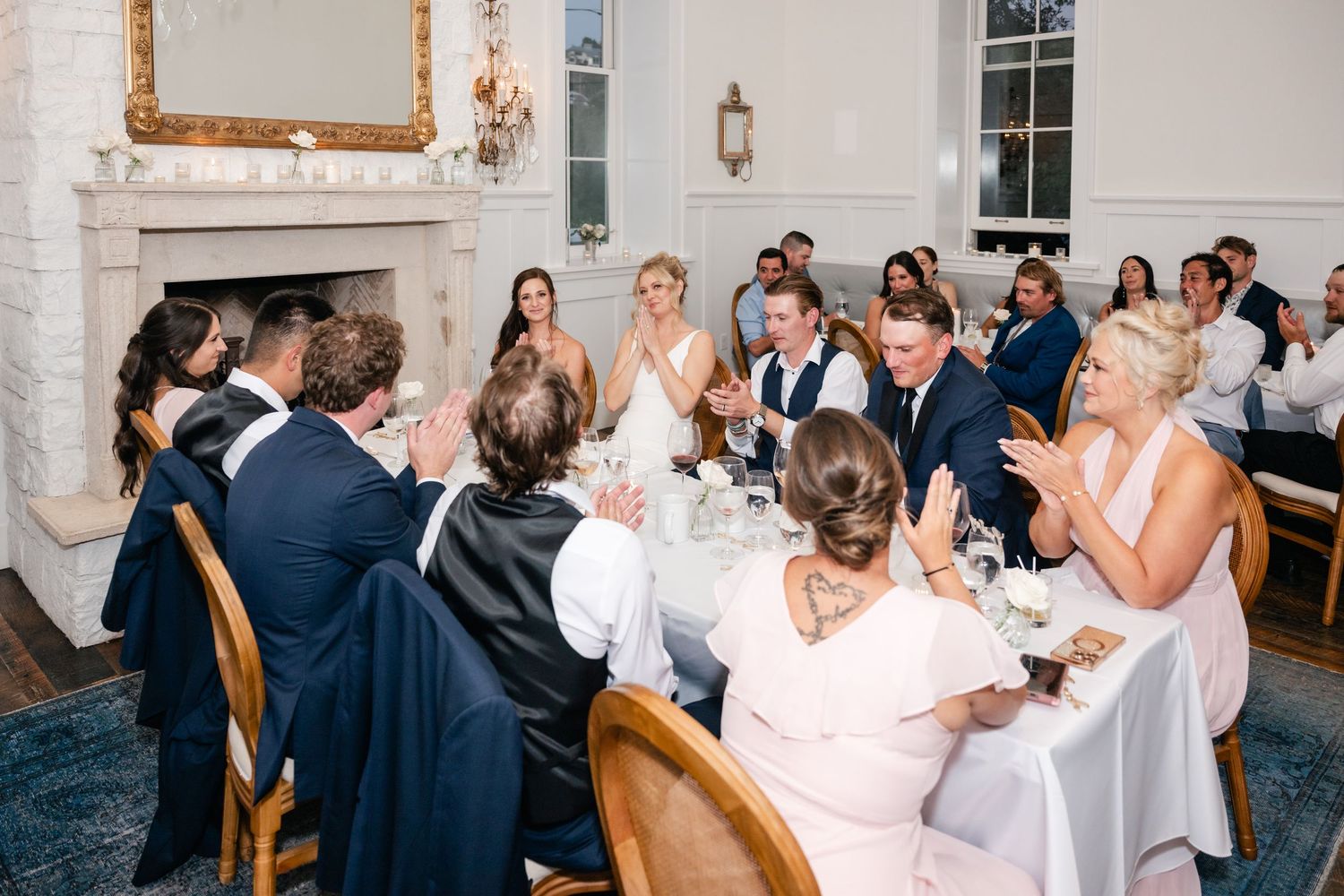 Wedding guests celebrate at round tables in an elegant white reception room with chandeliers.