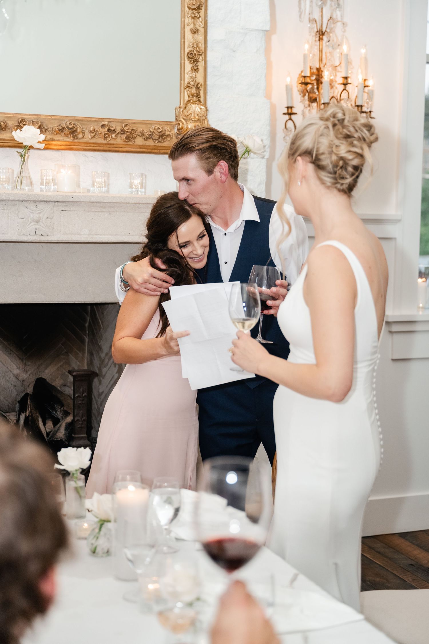 Guests sharing an emotional moment during wedding reception in an elegant white room with fireplace.