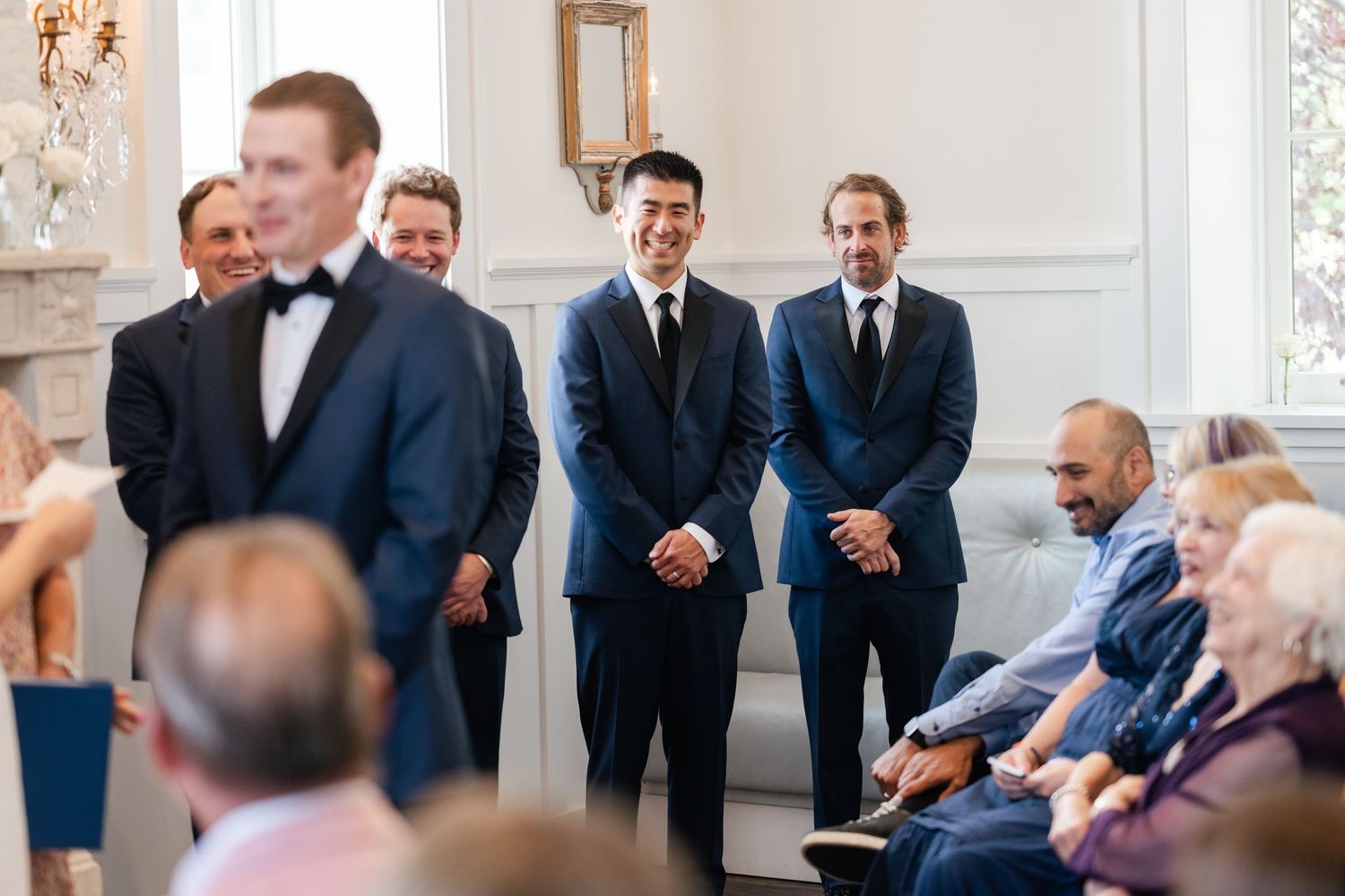 Wedding party members in formal attire stand in an elegant white room with chandelier.