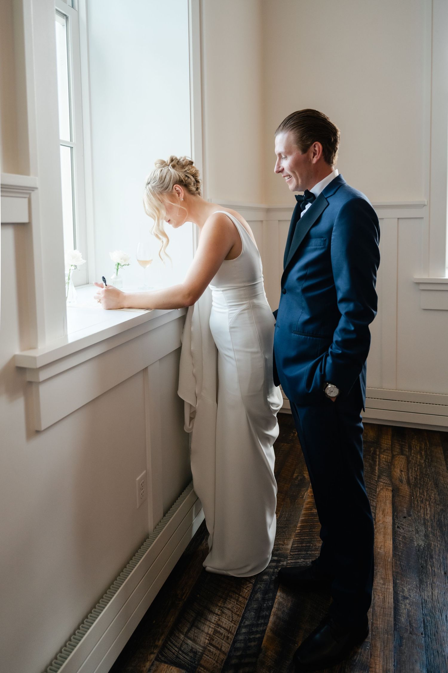 A couple shares an intimate moment by a bright window in a white-walled room with hardwood floors.