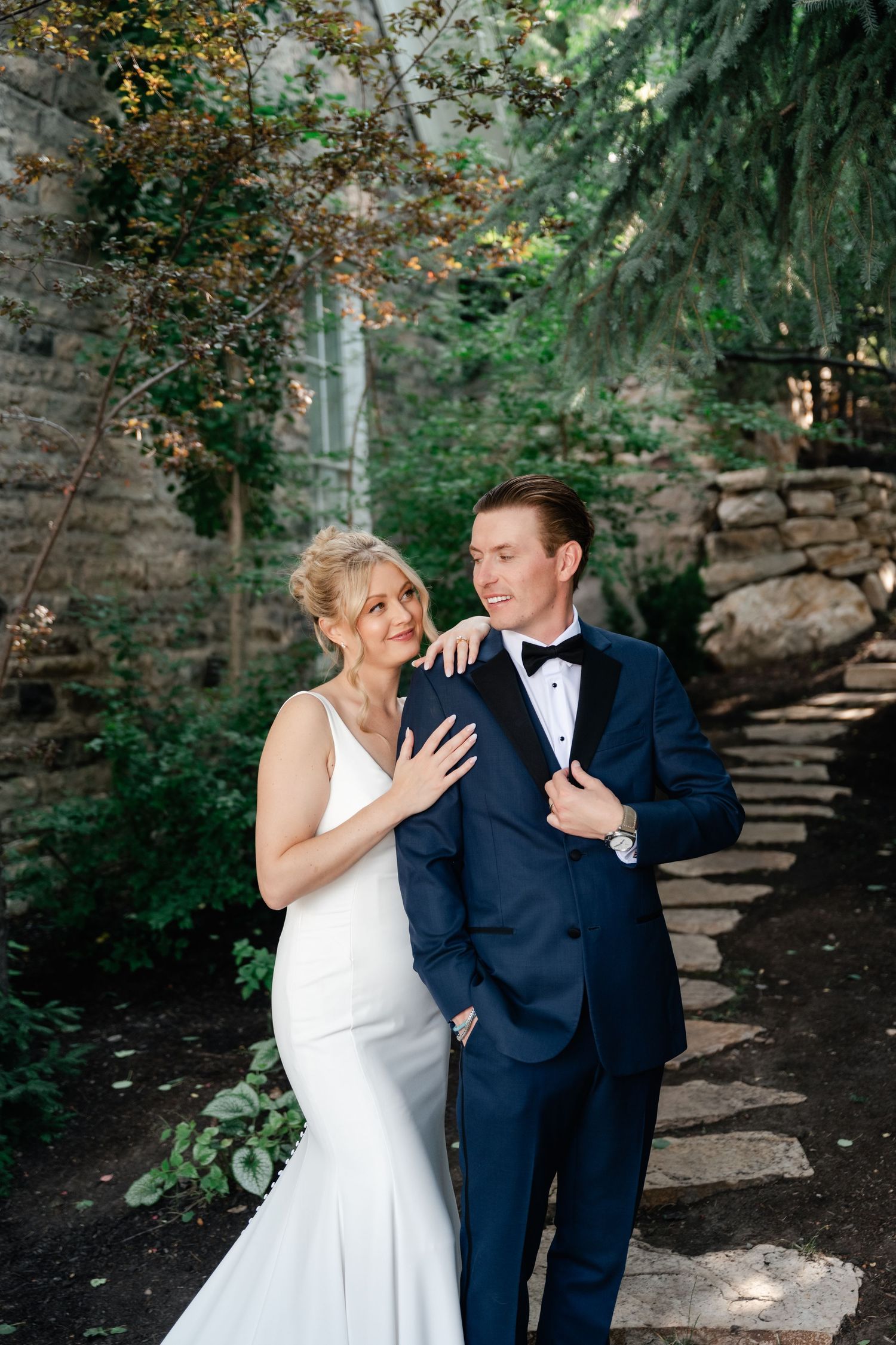 A joyful wedding couple shares a laugh together in a garden setting with stone walls.