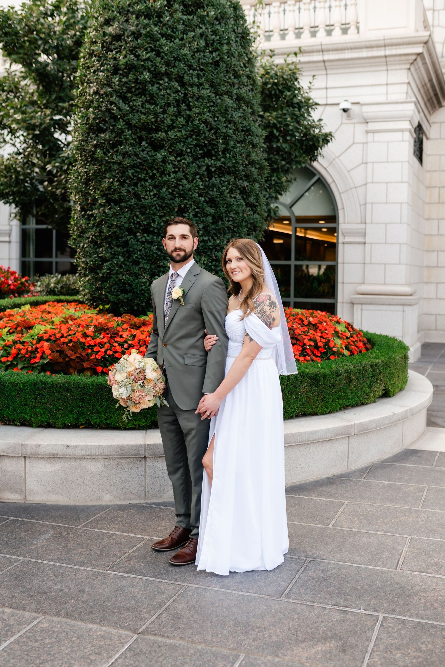 A couple poses in formal attire near blooming red flowers and greenery outside an elegant stone building.