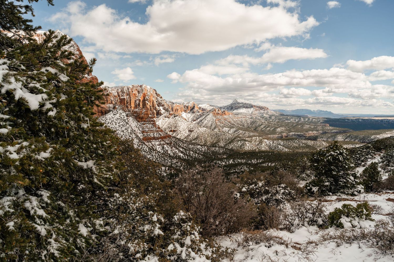 Panoramic winter landscape of snow-dusted red rock cliffs against a blue sky with white clouds.