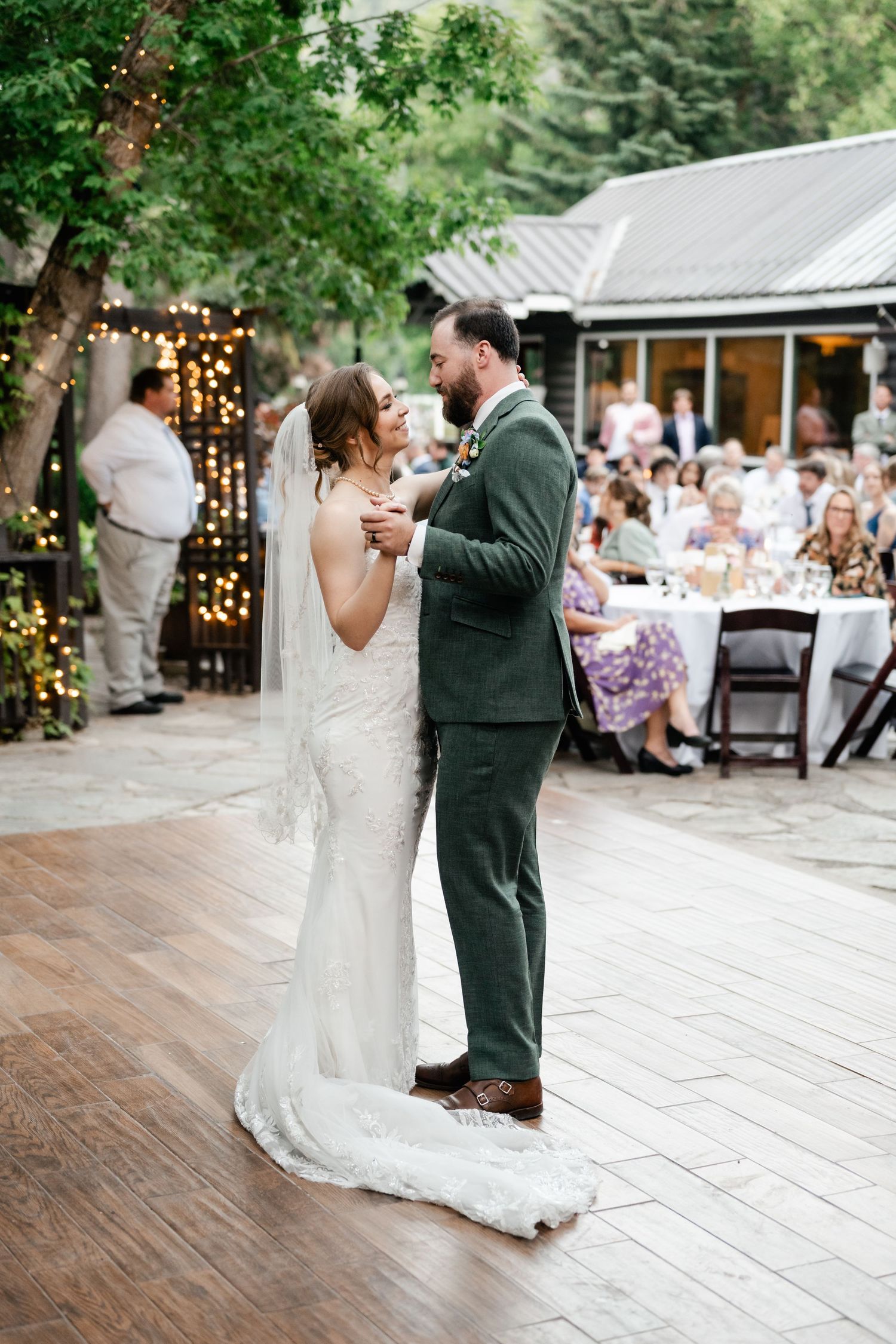 A couple shares their first dance on a rustic wooden deck during an outdoor wedding reception with string lights. Utah Wedding Photographer.