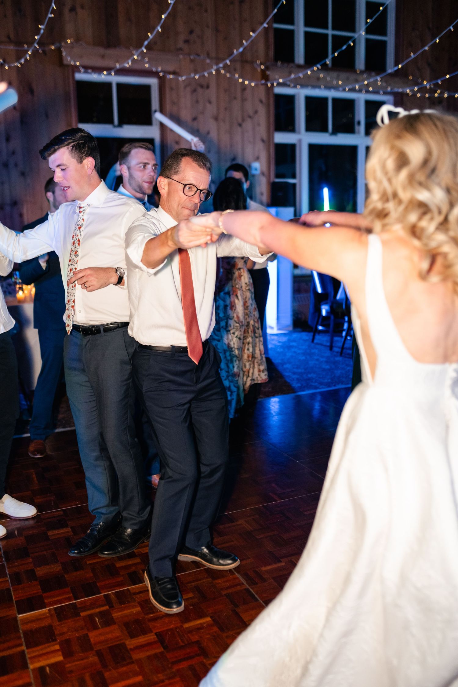 Wedding guests dance together at a rustic barn reception venue with string lights and wooden floors.