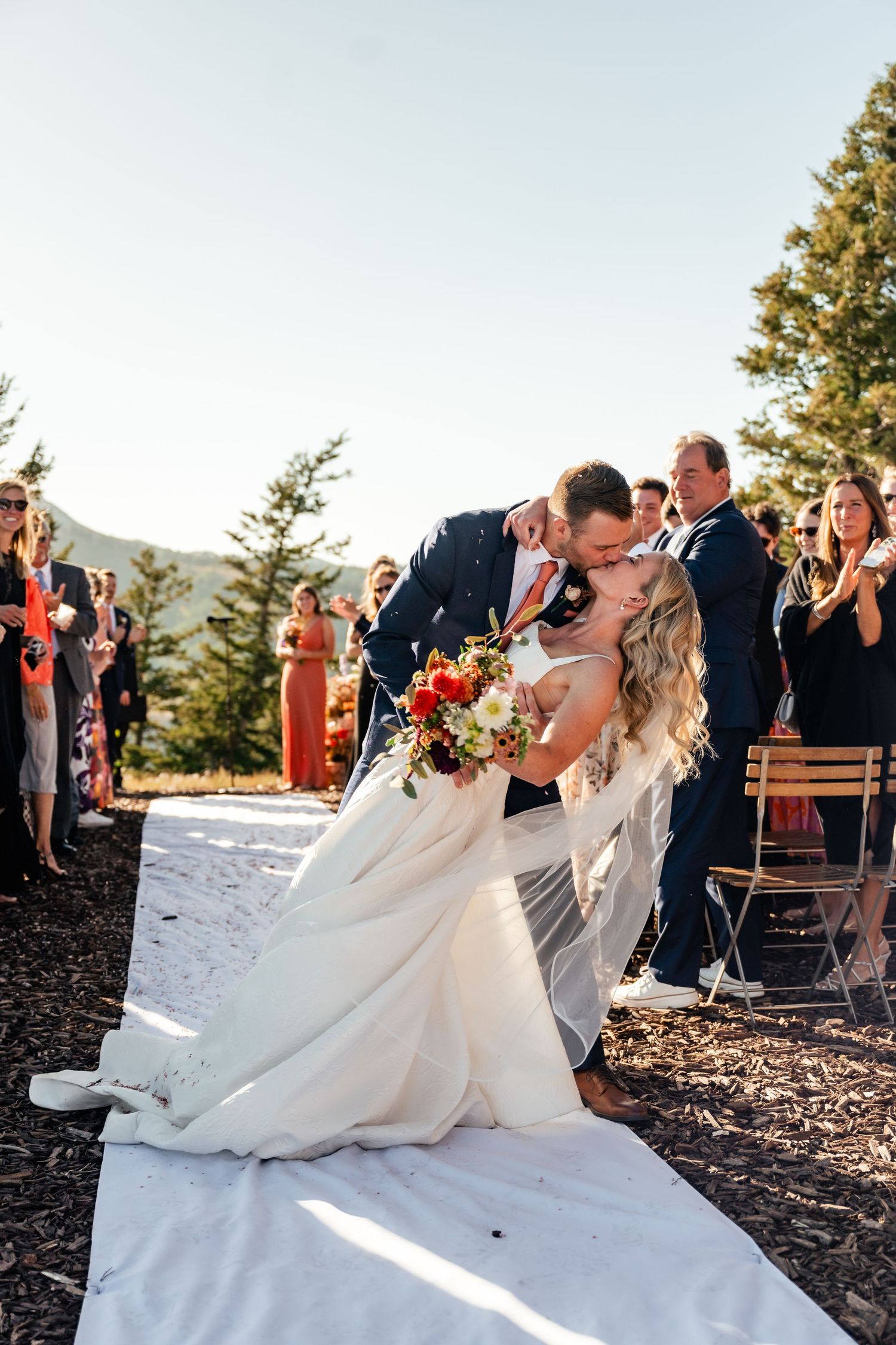 Wedding couple sharing first kiss at mountain ceremony while guests applaud.