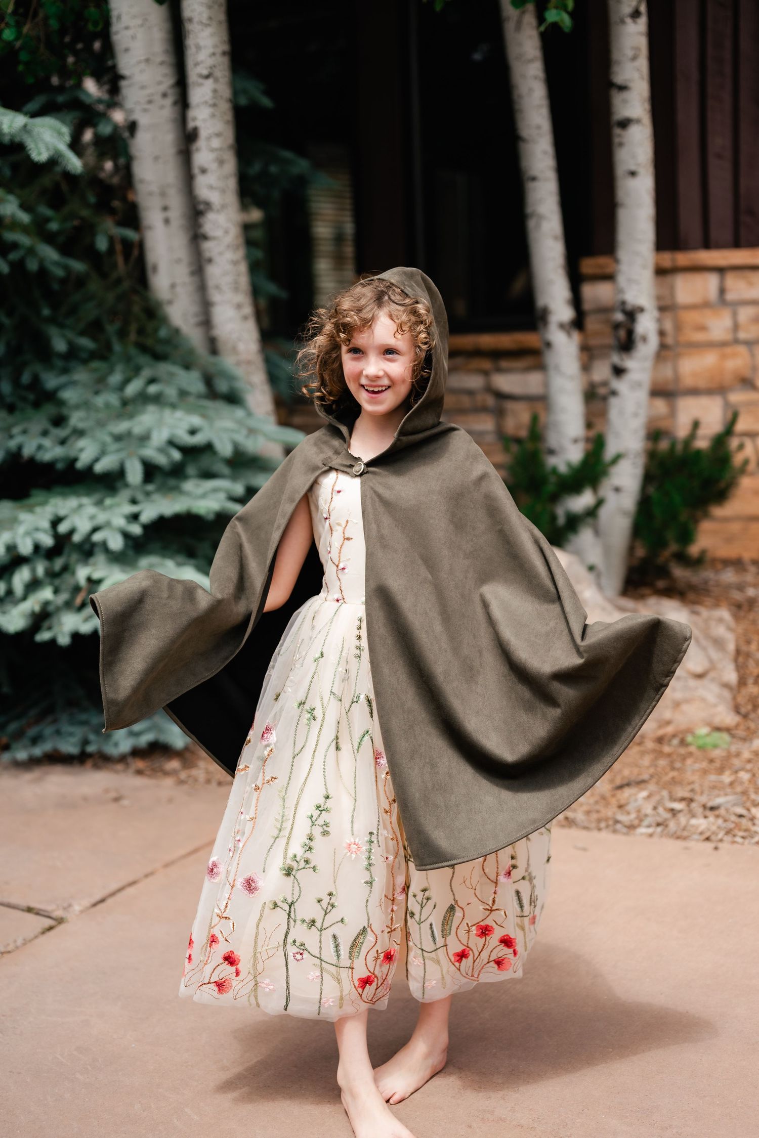 Child wearing floral dress and gray cape twirling outdoors near birch trees.