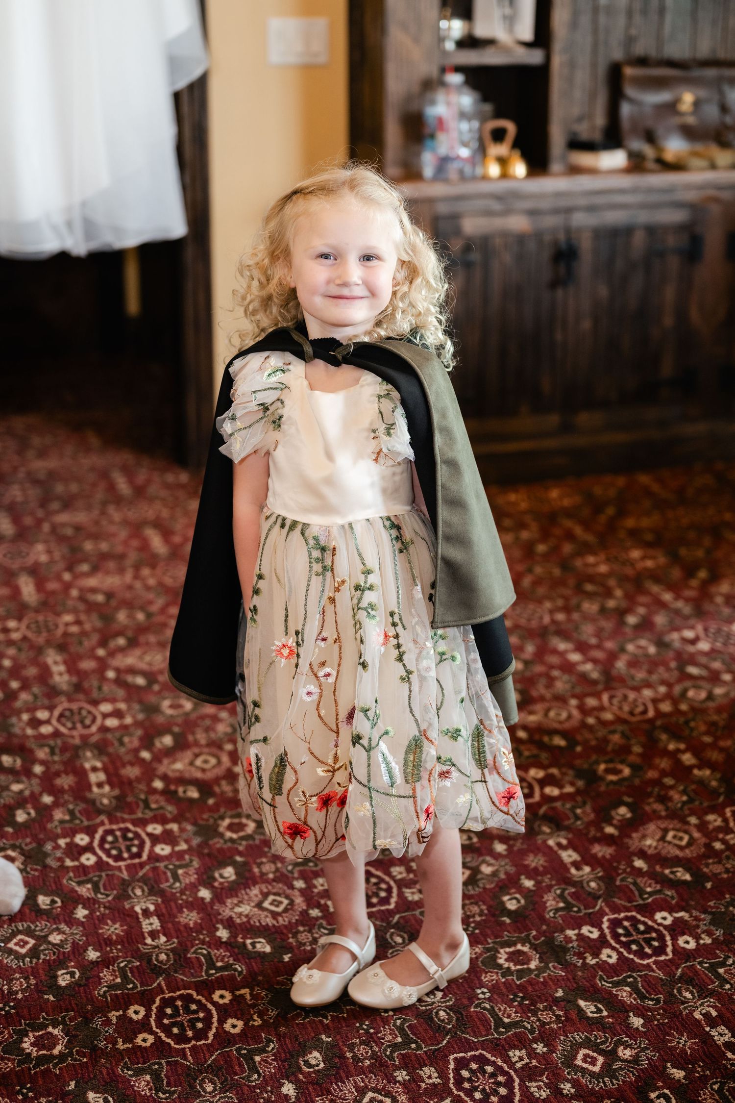 A small child in a floral dress smiles while standing on a patterned carpet.