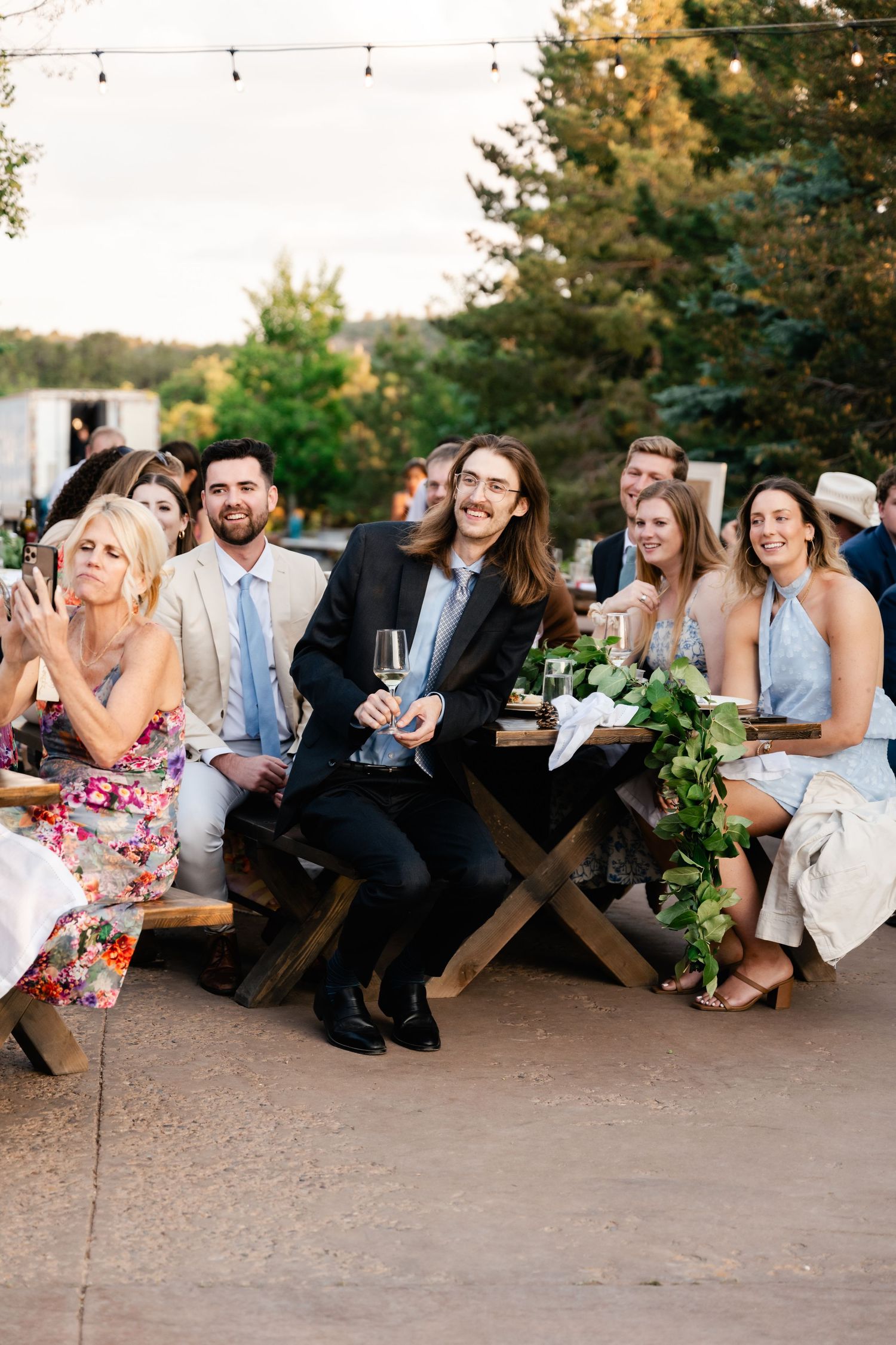 Wedding guests seated on wooden benches at an outdoor reception share laughter and smiles.