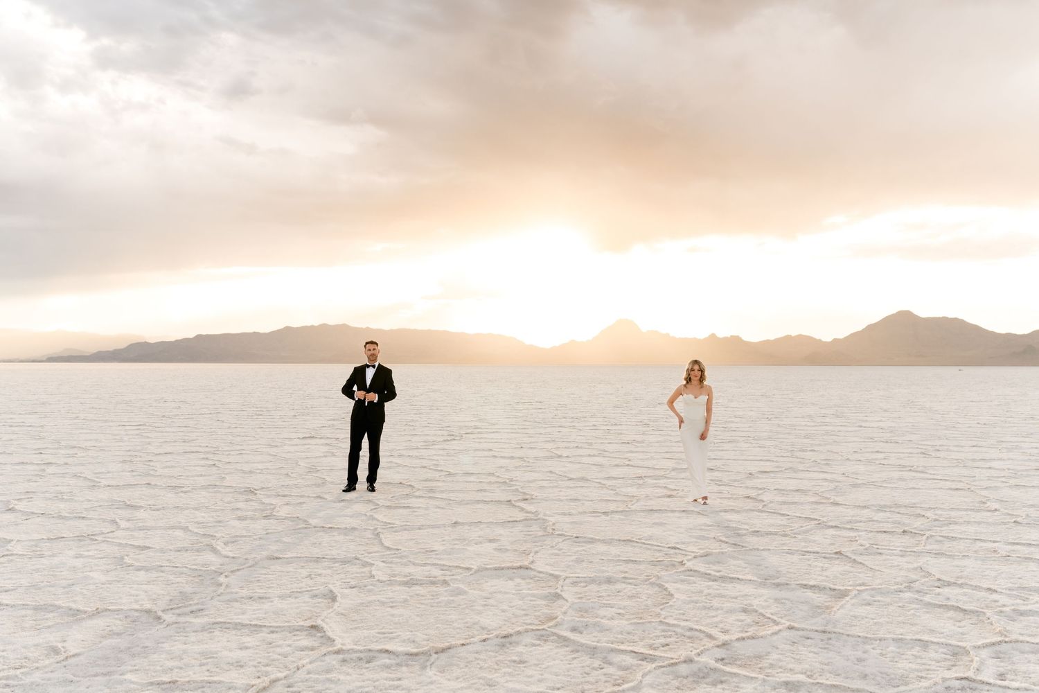 A panoramic couple portrait at vast salt flat landscape at sunset with distant mountains and silhouetted figures. Salt Flats Photographer.