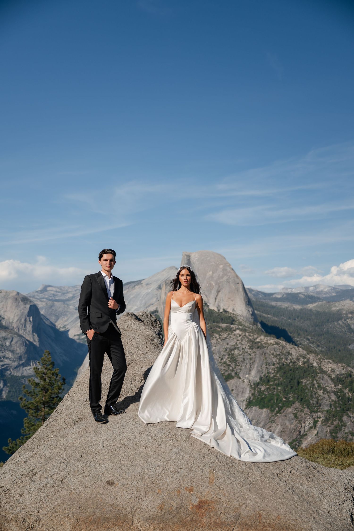 Bride and groom pose on a rocky summit with Half Dome and Yosemite Valley in the background on their wedding day.