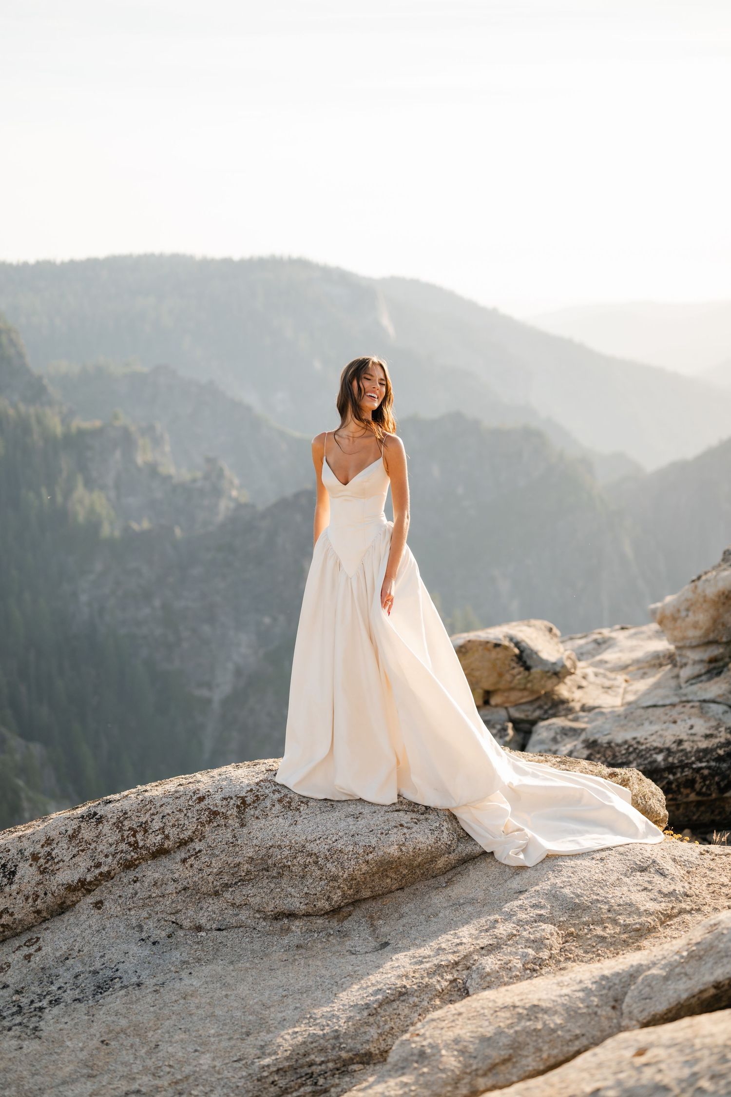Bride in flowing white gown stands on rocky mountain cliff with misty peaks backdrop.