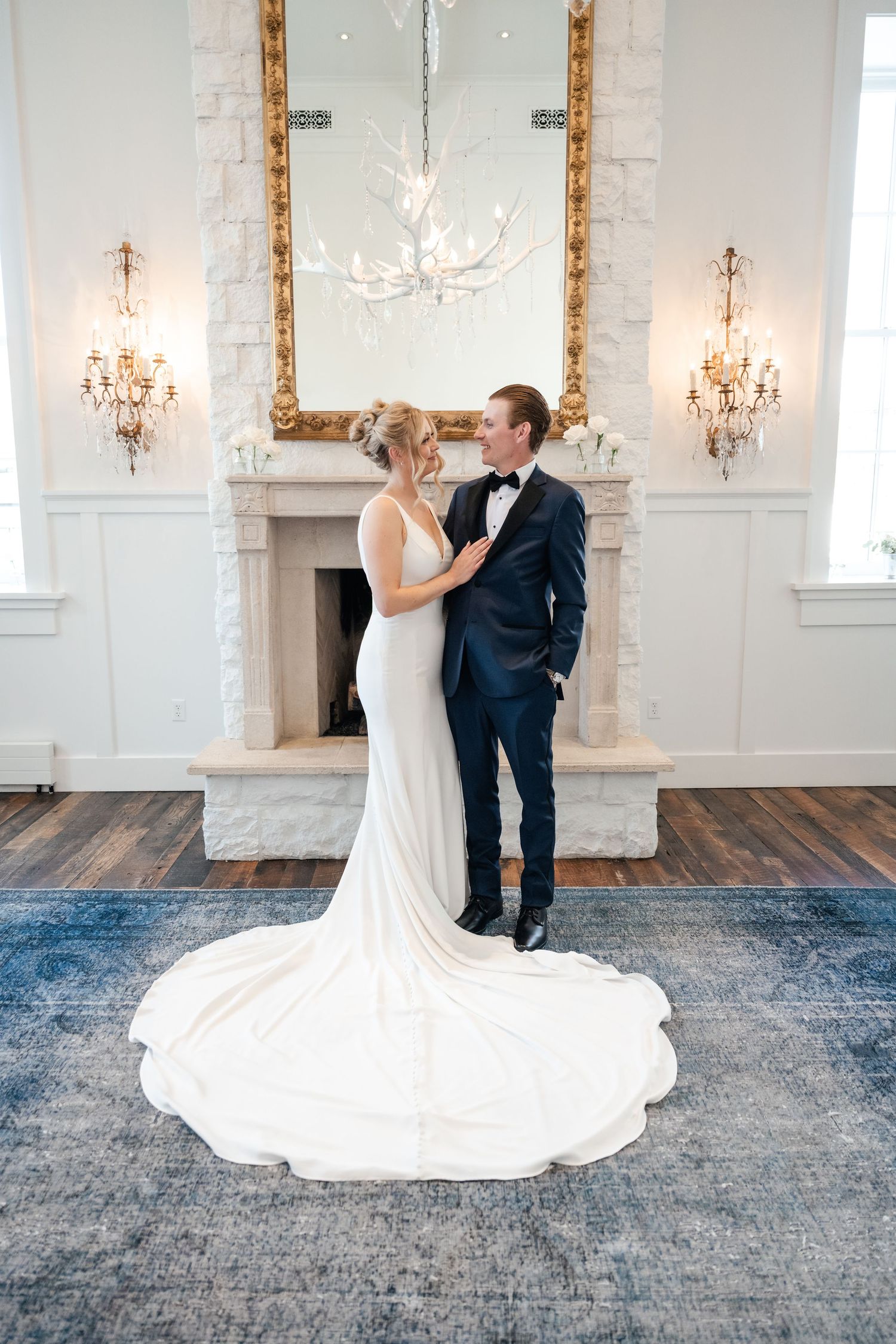 Bride in elegant white gown with train stands with groom in navy tuxedo by ornate fireplace and mirror of the Washington School House House.