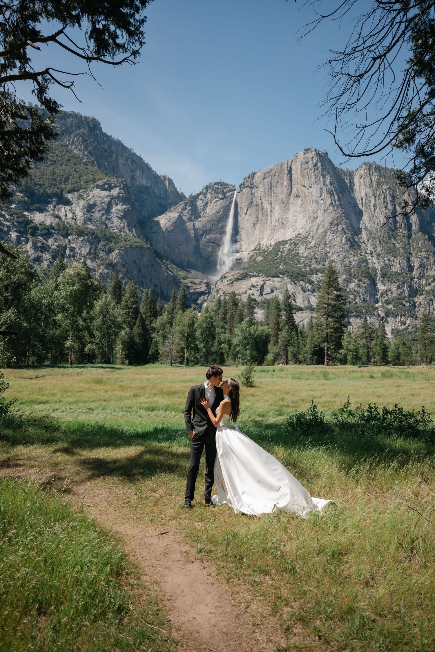 Wedding couple shares romantic kiss on trail in Yosemite Valley with stunning waterfall behind them.