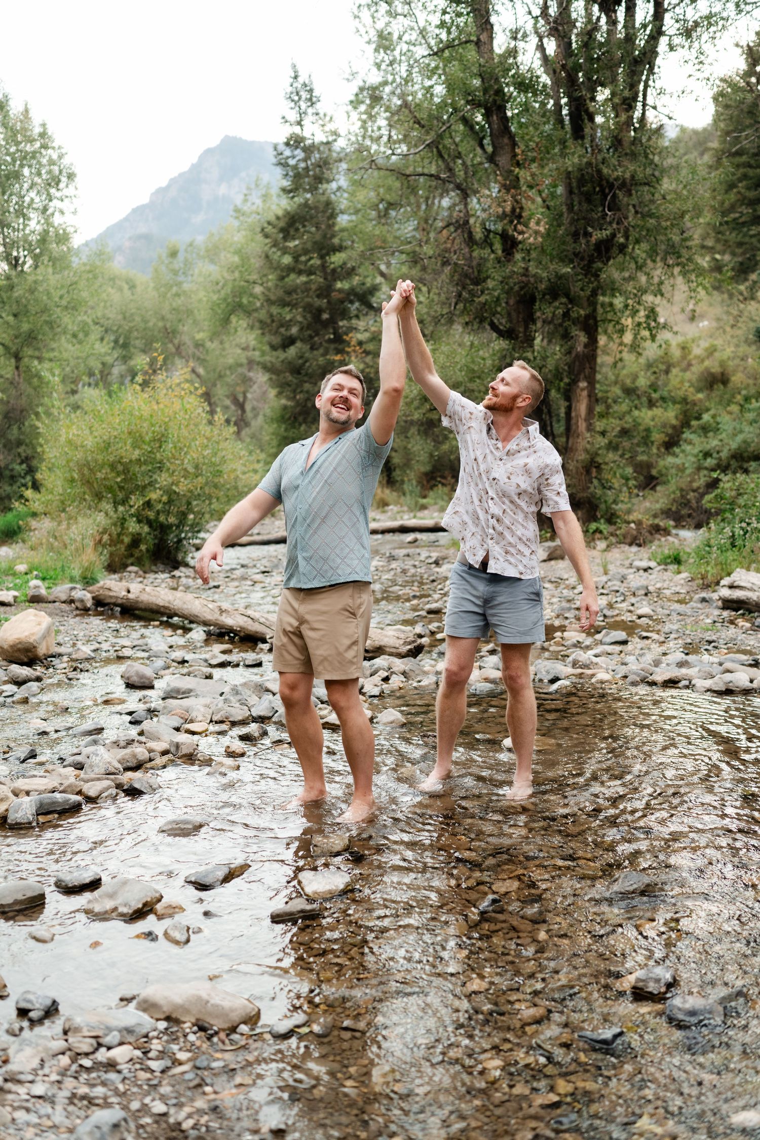 LGBTQ couple engagement photos in the Utah mountains. Two men laugh and hold hands while wading barefoot through a rocky mountain stream.