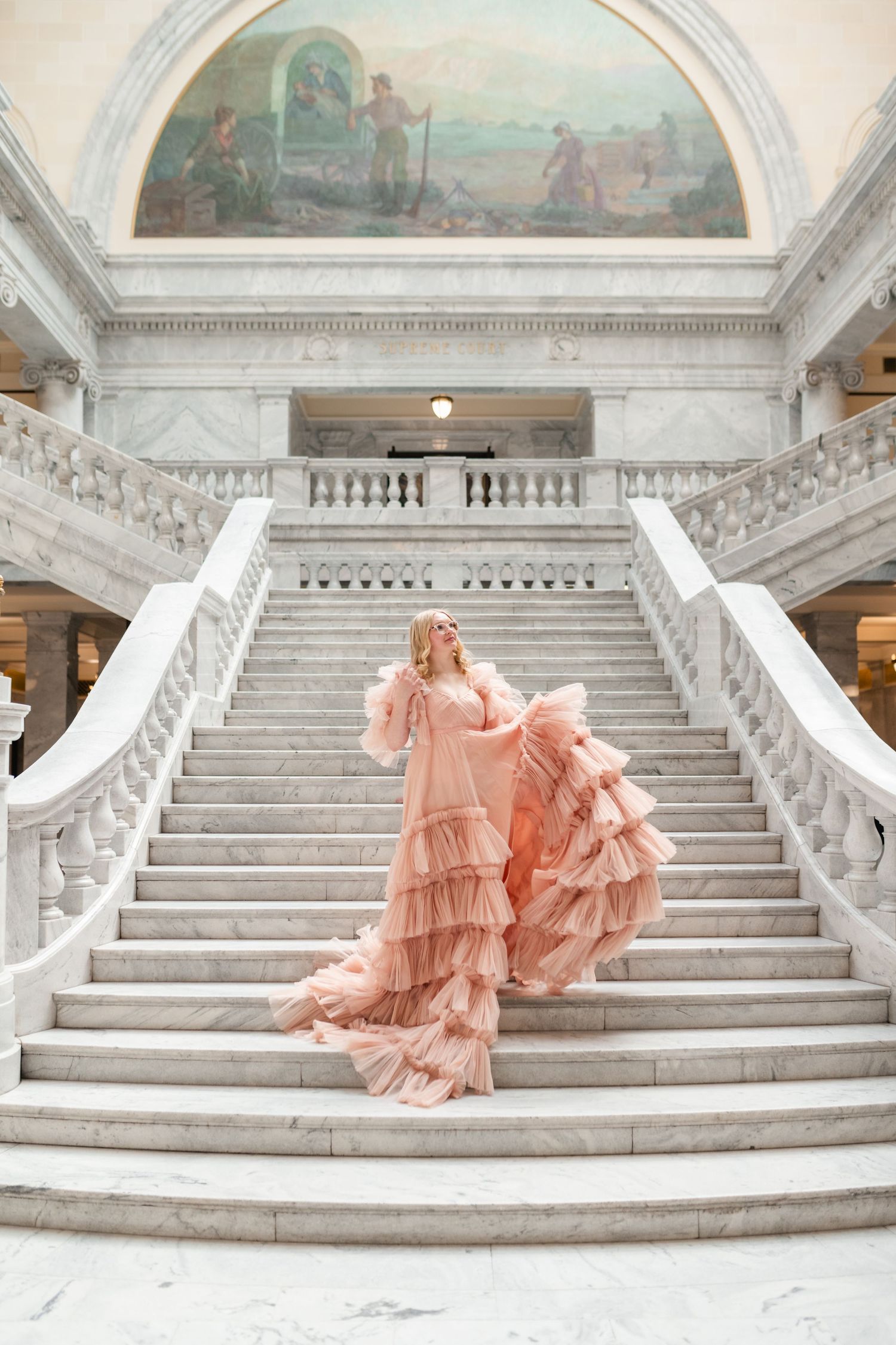 A romantic pink dress flows across white marble steps beneath arched ceilings adorned with classical murals.